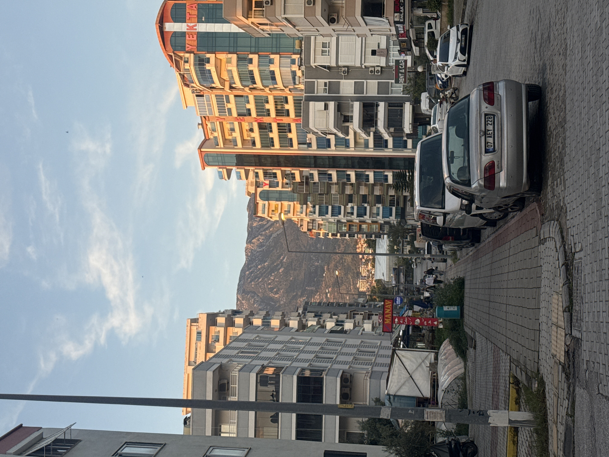 Here is a detailed description of the image:
The image depicts a street scene in a city, with a mountain range visible in the distance. The street is lined with buildings of varying heights and architectural styles. The buildings on the left side of the street are primarily gray and white, while the buildings on the right side are predominantly beige and orange. There is a mix of residential and commercial buildings, indicated by the presence of balconies and storefronts.
Cars are parked along the side of the street, and there are some pedestrians visible in the distance. A sign with the word "MANAV" is visible on the lower right side of the image. The sky is blue with some scattered clouds. The road is paved with bricks.