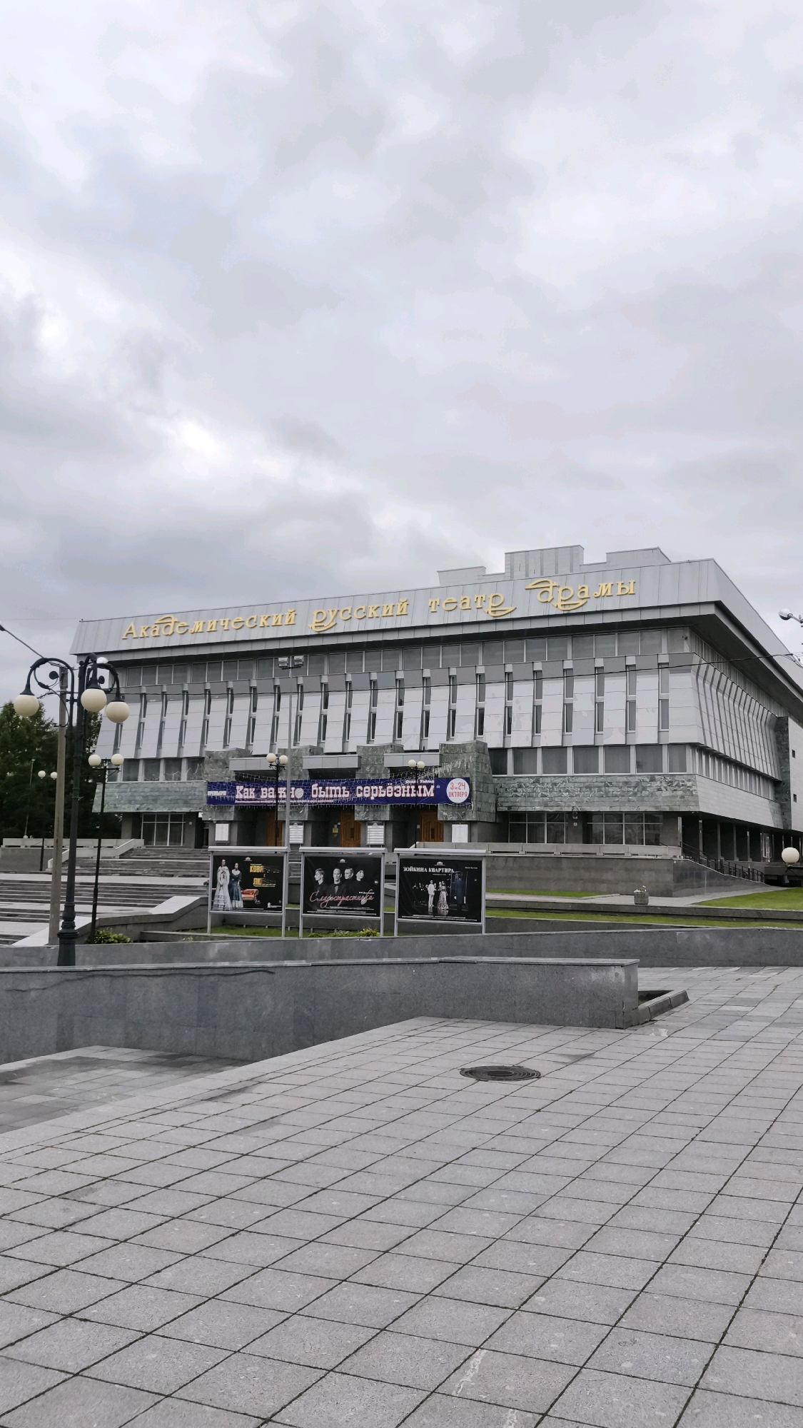 The image shows the Academic Russian Drama Theater in Yoshkar-Ola, Russia. The theater is a large, modern building with a grey facade and golden lettering that reads "Академический Русский театр драмы" (Academic Russian Drama Theater). The building has a prominent entrance with stairs leading up to it. In front of the theater, there are several posters displayed on stands, advertising upcoming performances. One poster features text that appears to be a play title. The sky is overcast. In the foreground, there is a paved area with a patterned walkway and several lampposts.