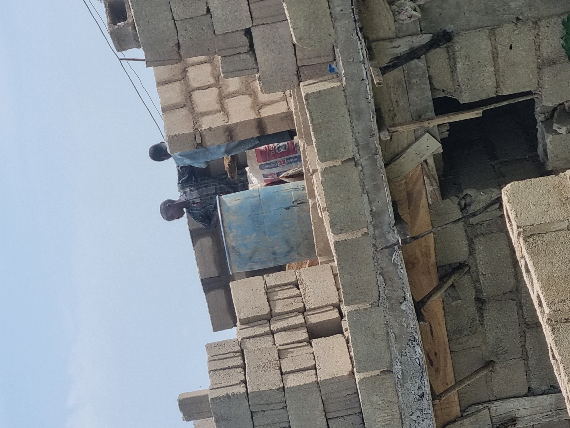 The image depicts a construction site in Jos, Nigeria, with two individuals working on a building. The foreground shows stacked concrete blocks, forming the walls of the structure. A large blue barrel is visible on the right side of the image, possibly used for storing materials. Behind the barrel, a bag of cement is leaning against the wall. The two men are positioned on the upper level of the building, surrounded by more concrete blocks. One man, wearing a checkered shirt, appears to be looking downwards, while the other, in a light blue garment, is facing away from the camera. The sky above is clear and pale blue. The scene suggests ongoing construction work, with materials and people engaged in the building process.
