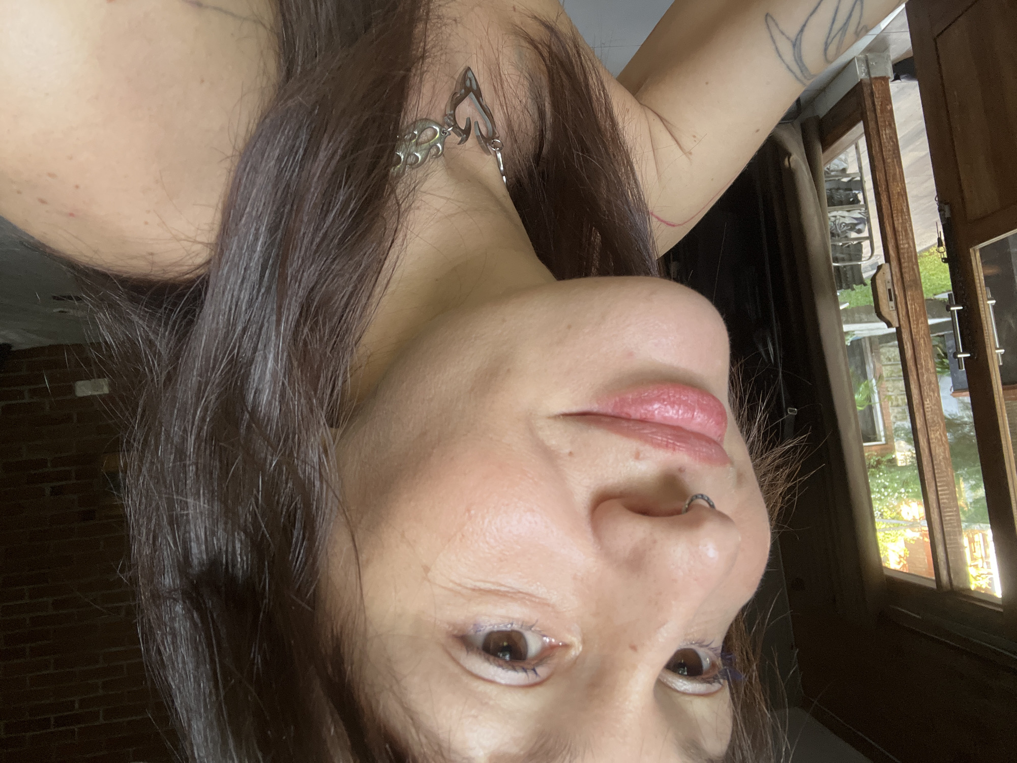 A portrait of a woman is taken indoors from a low angle, making her look down at the camera. The woman has long, dark hair and fair skin. She wears a metal necklace with a heart-shaped pendant and a nose ring. Her left arm has a tattoo. A window is behind the woman. Outside the window, there is greenery. The location in the city of North Kuta, Indonesia is unknown.