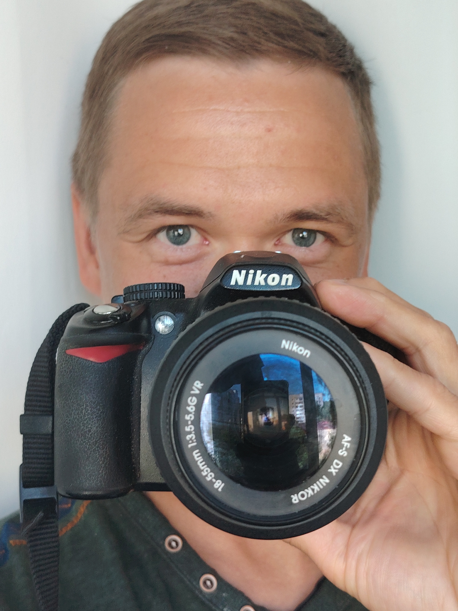 A man is taking a selfie with a Nikon camera. He is holding the camera up to his face, and only his eyes, forehead, and a portion of his hair are visible above the camera. The camera lens reflects a building and what appears to be a person. He is wearing a dark green shirt with buttons. The background is a plain, light-colored wall. The image was taken in Chaikovsky, Russia.