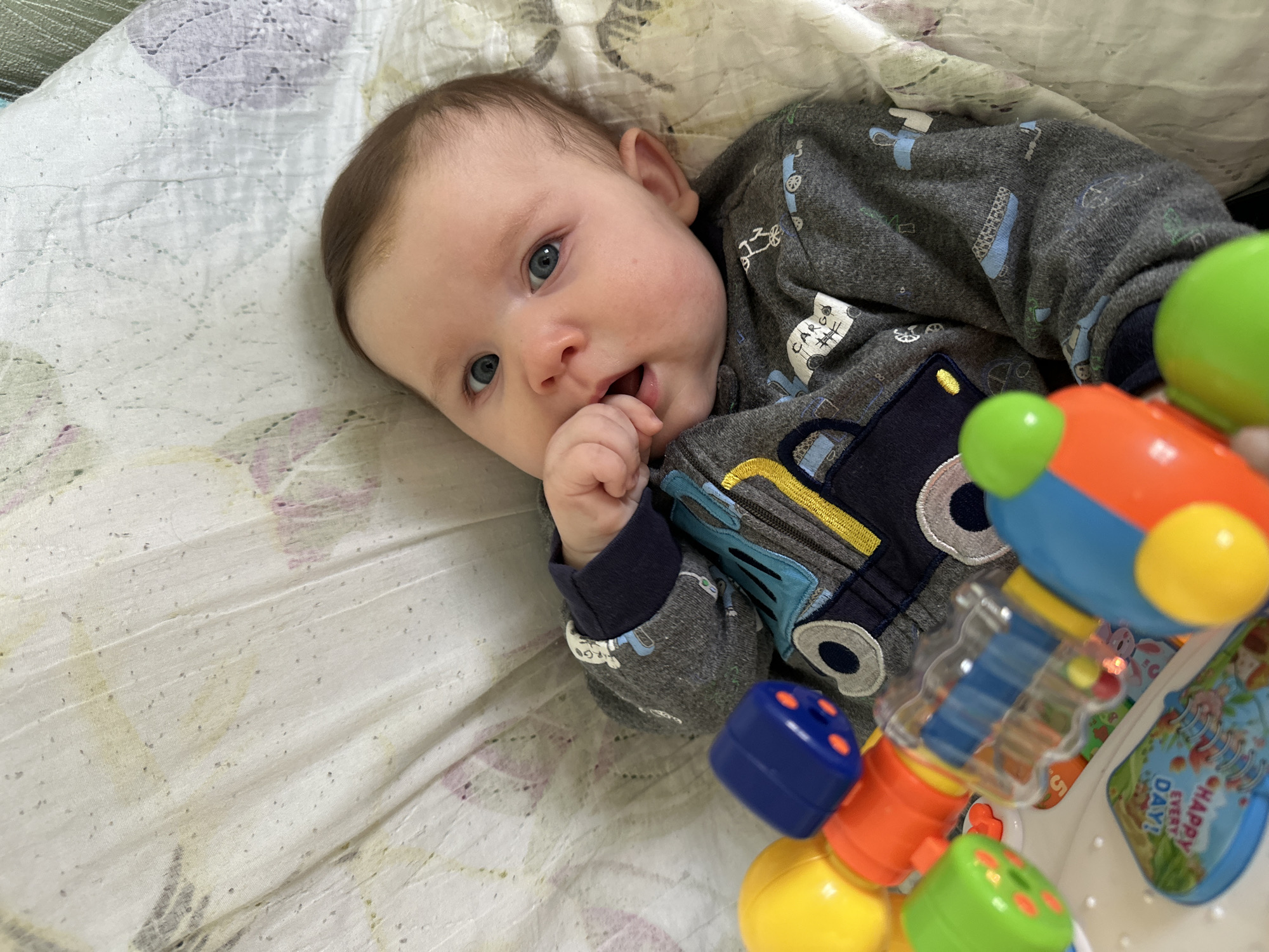 An infant lies on their back on a light-colored, subtly patterned blanket or bedding, looking directly towards the camera. The baby has fair skin, bright blue eyes, and light brown hair, with one hand held up near their open mouth. They appear calm and curious.
The baby is dressed in a grey long-sleeved onesie decorated with an all-over pattern of small blue and white vehicles, including trucks and cars. A larger, dark blue and yellow truck appliqué is prominent on the chest of the onesie. Visible text on the smaller vehicle patterns includes "CARGO".
In the bottom right foreground, a colorful plastic toy is partially visible, featuring bright blue, orange, yellow, and green components, some with clear plastic sections, suggesting it might be a stacking toy or rattle. Part of the toy or an item behind it displays the text "HAPPY EVERY DAY!" on a blue background with illustrations.
The scene is indoors, with soft, even lighting. The overall impression is one of a peaceful moment. While the location context is Chaikovsky, Russia, no specific visual cues in the image confirm this.