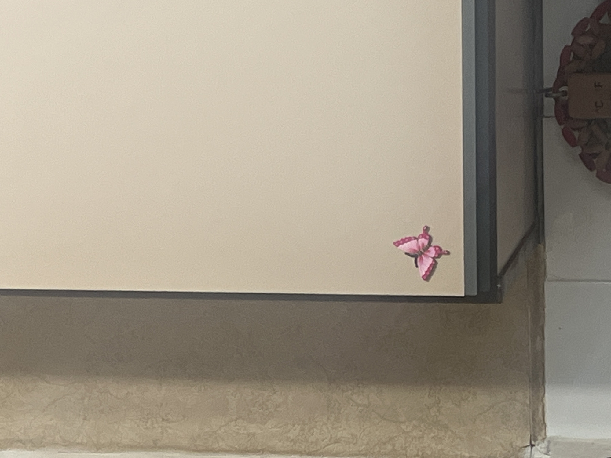 The image presents an indoor scene located in Burgas, Bulgaria. A light beige rectangular surface, likely a cabinet door or panel, dominates the right side of the frame. A vibrant pink decorative butterfly, possibly a decal or sticker, is affixed to the lower-left section of this light-colored surface. To the left, a textured wall, appearing to be covered in patterned wallpaper or painted plaster, is visible in tones of beige and light brown, showing minor discoloration and wear at its base. A distinct dark vertical line separates this textured wall from the adjacent light-colored surface. Below the light-colored surface, a dark horizontal strip indicates the base of a structure, possibly a shelf or the underside of the cabinet. Partially visible below this strip is a decorative object, possibly a wreath, from which a light-colored tag with faint markings, potentially "C F," is suspended. The lower-left corner reveals a white surface, possibly tiled or painted, with a noticeable seam or grout line showing slight dirt accumulation. No individuals, explicit actions, or interactions are depicted within the scene.