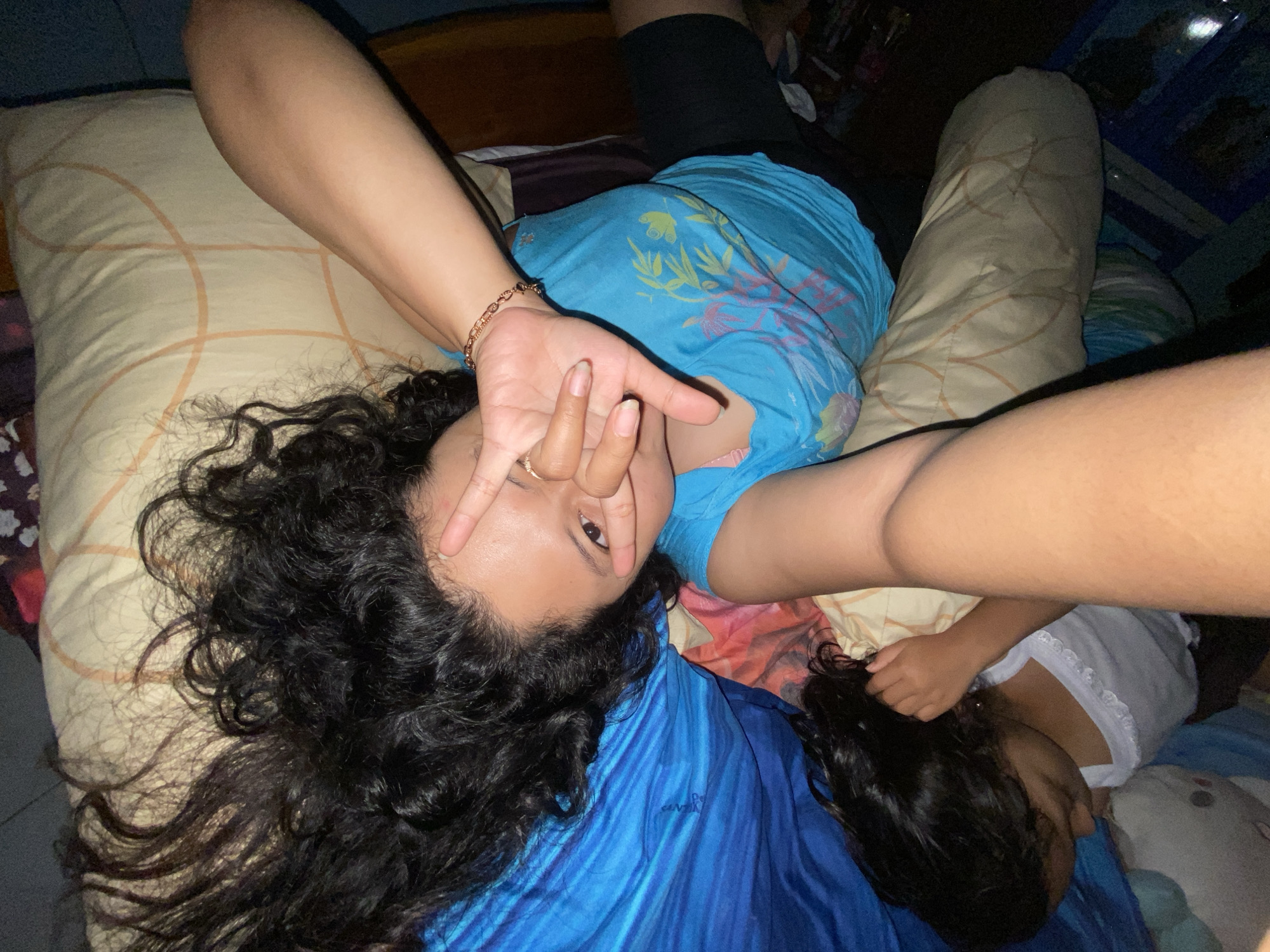 In this overhead shot, two people are lying in bed in Gelam, Indonesia, at night. The subject in the foreground, facing the camera, has long, dark, curly hair. She is wearing a blue t-shirt with a floral design and black shorts. Her left arm is raised, with her hand near her face, obscuring part of it. Her fingers form a peace sign gesture over her eye. She is also wearing a ring on her right ring finger and a bracelet on her left wrist.
Beside her, a younger child is lying on their side, facing away from the camera. The child has dark hair and is wearing a white t-shirt with lace trim. The bed is covered with light-colored bedding, and there is a blue blanket or sheet in the foreground. The background is dark, suggesting nighttime. The lighting is dim, and no specific weather cues are visible. The overall mood appears casual and relaxed.