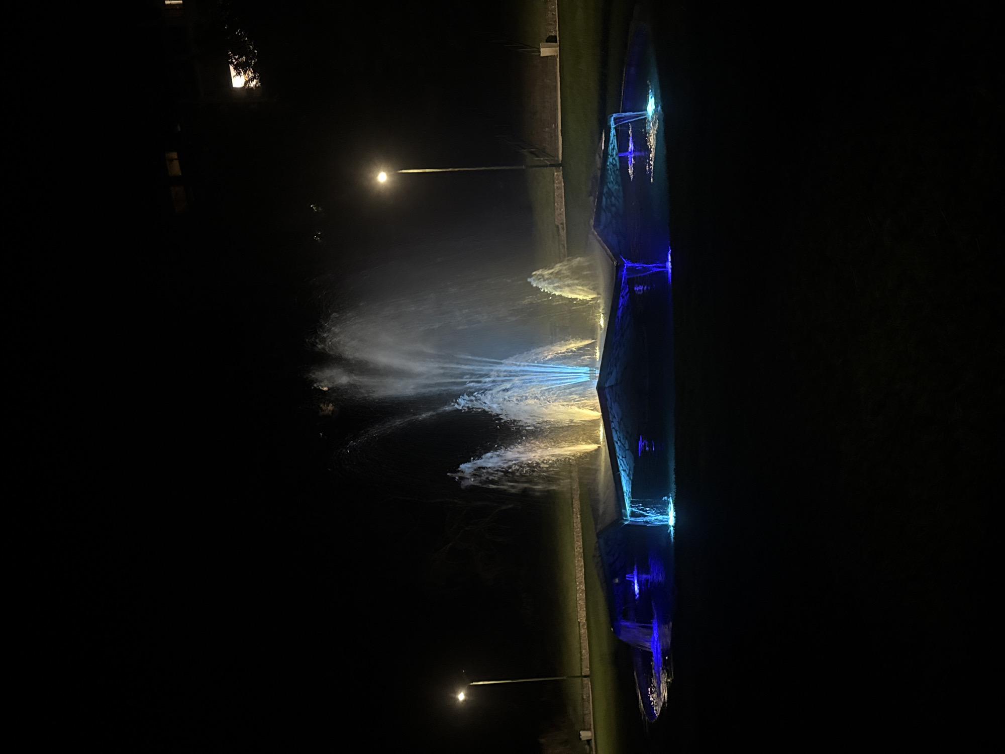 A nighttime, medium shot captures a public fountain illuminated by blue and yellow lights, located in the city of Sandanski, Bulgaria. The fountain is the central subject, with water jets reaching upward and cascading into a lower pool. The surrounding area is dark, with a lamppost to the left providing a faint glow and a building visible in the upper right corner. The ground is covered in grass. No people are present in the frame. The scene depicts a decorative public amenity at night.