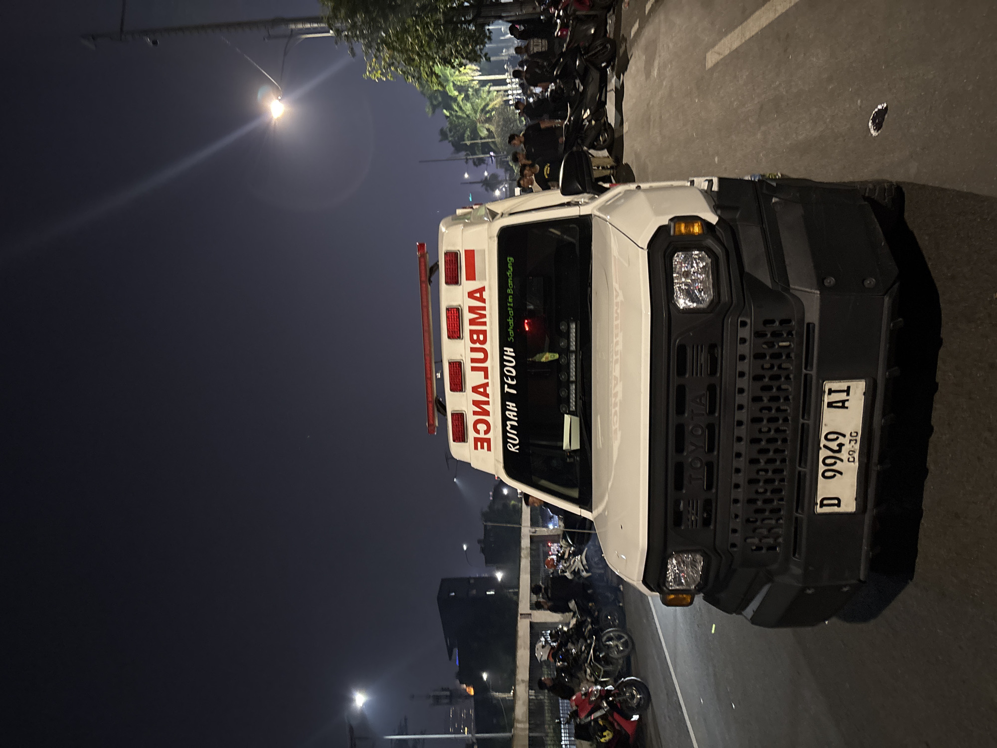 An ambulance is parked on an asphalt road at night, possibly in Central Jakarta, Indonesia. The ambulance is white with red emergency lights on top and the word "AMBULANCE" written in red across the front. The text "RUMAH TEDUH Sahabat di Bandung" is visible on the windshield. A license plate reading "D 9949 AI" is mounted on the front bumper. Several people are standing around the ambulance and on the side of the road. Motorcycles are parked on the left side of the ambulance. In the background, buildings and streetlights are visible, indicating an urban environment. The scene appears to be calm and orderly.