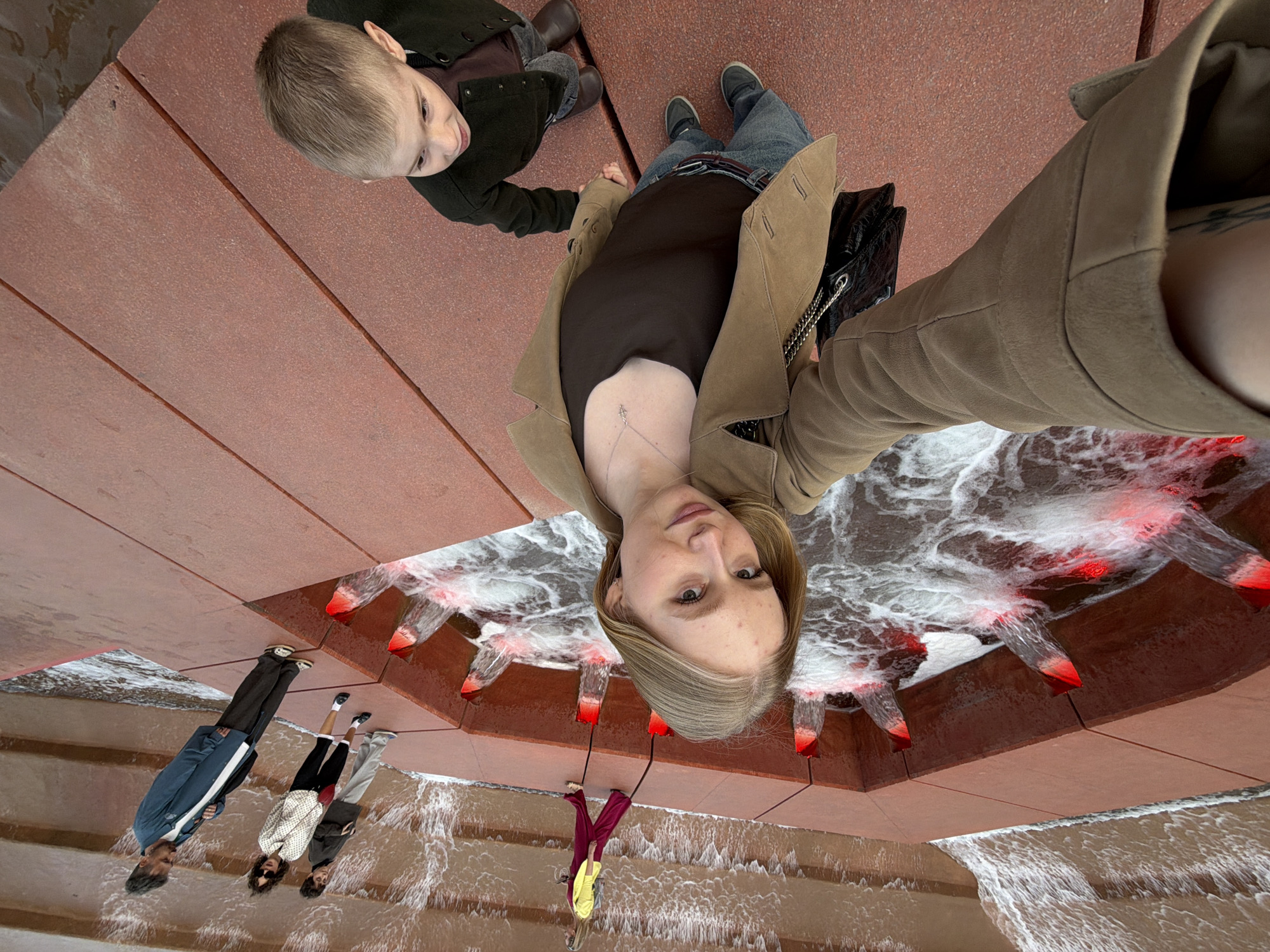 A low-angle shot captures a woman and a young boy standing next to a fountain, taking a selfie. The boy, in a dark jacket and brown pants, holds the woman's hand. The woman wears a tan coat, a black top, and jeans. The fountain has red accents on the water outlets. The background includes other people near the fountain's edge, and the overall setting resembles an urban public space. The image is oriented upside down. There is no explicit indication this photo was taken in Krasnodar, Russia.