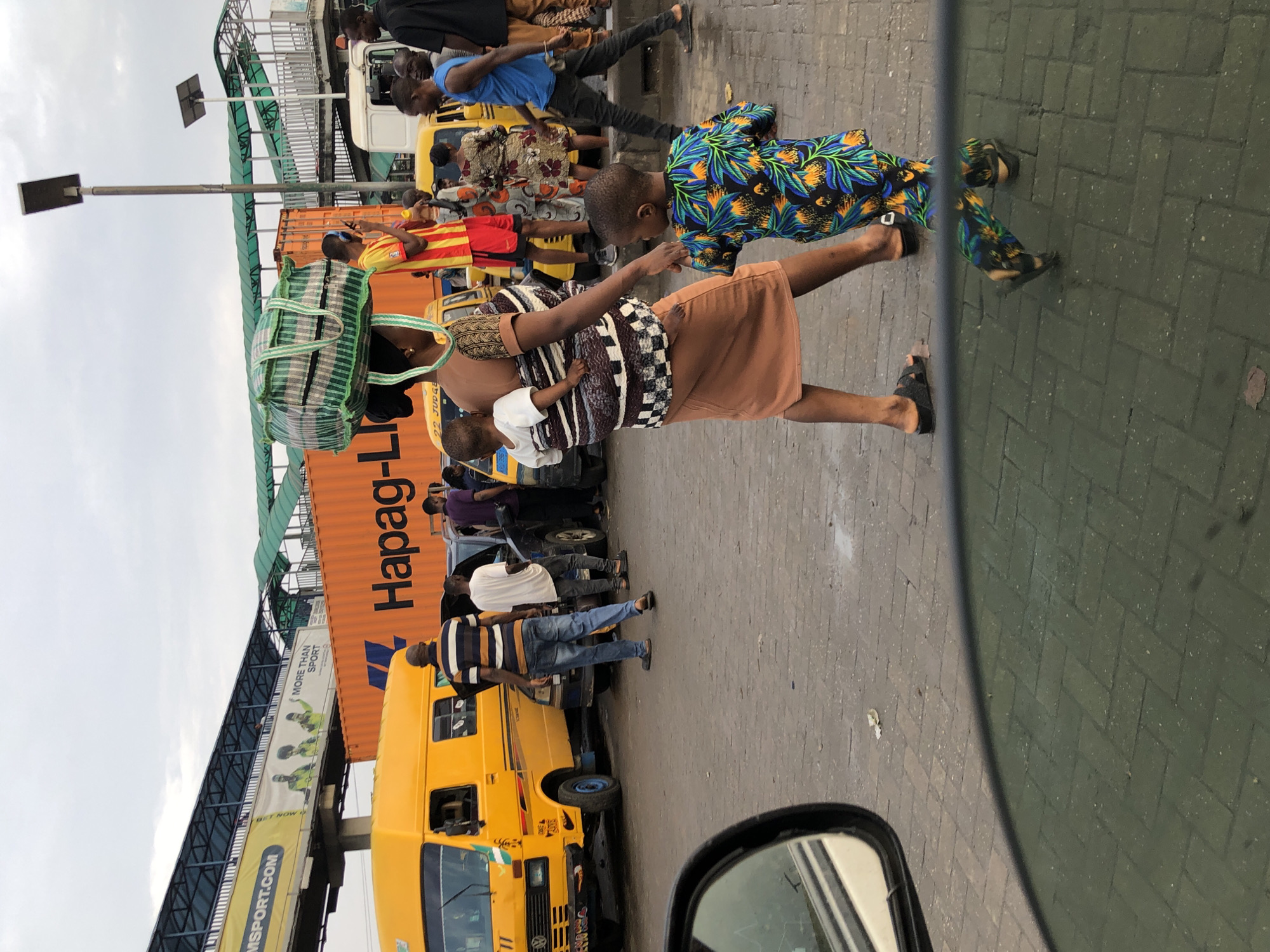 The image depicts a busy street scene in Ikeja, Nigeria, likely near a transport hub given the presence of a yellow bus and a large shipping container. A woman, carrying a large bundle of goods on her head, walks with two young children. One child is strapped to her back, and the other walks beside her, holding her hand. She is dressed in traditional clothing, with a patterned fabric wrapped around her torso and a brown skirt. The child beside her wears a vibrant, floral-patterned shirt.
In the background, a yellow Volkswagen bus is parked, its hood open. Several people are visible around it, including a man in a striped shirt and jeans, and another in a white t-shirt. Further back, a large orange shipping container with "Hapag-Lloyd" branding is partially visible. Another yellow vehicle, possibly a taxi or a bus, is seen to the right, with several passengers. The environment suggests a commercial or transportation area, with overhead structures that could be part of a pedestrian bridge or an elevated road. The ground is paved with interlocking blocks, and the sky is overcast, indicating a possibly humid climate. The scene is captured from a low angle, possibly from within another vehicle, as suggested by the reflection in the lower portion of the image.