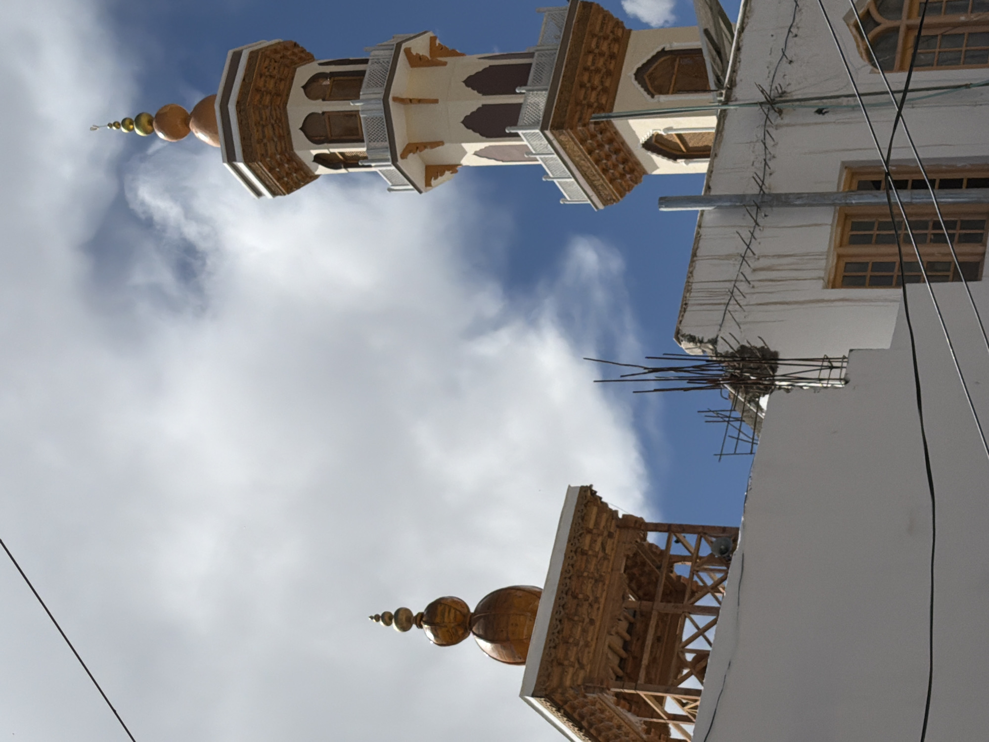 The image depicts a view of the Leh Palace minaret and a wooden stupa-like structure in Leh, India, against a backdrop of a cloudy sky. The minaret, a tall, slender tower with multiple levels and arched windows, is adorned with intricate wooden carvings and a golden finial. The wooden structure to the left features decorative carvings and a similar finial. The foreground shows the white walls of buildings, one with visible water stains, and several wooden-framed windows. Overhead, electrical wires crisscross the frame, and scaffolding with rebar is visible on one of the buildings, suggesting ongoing construction or repair. The overall scene captures a blend of architectural styles and the ambient conditions of a high-altitude location.