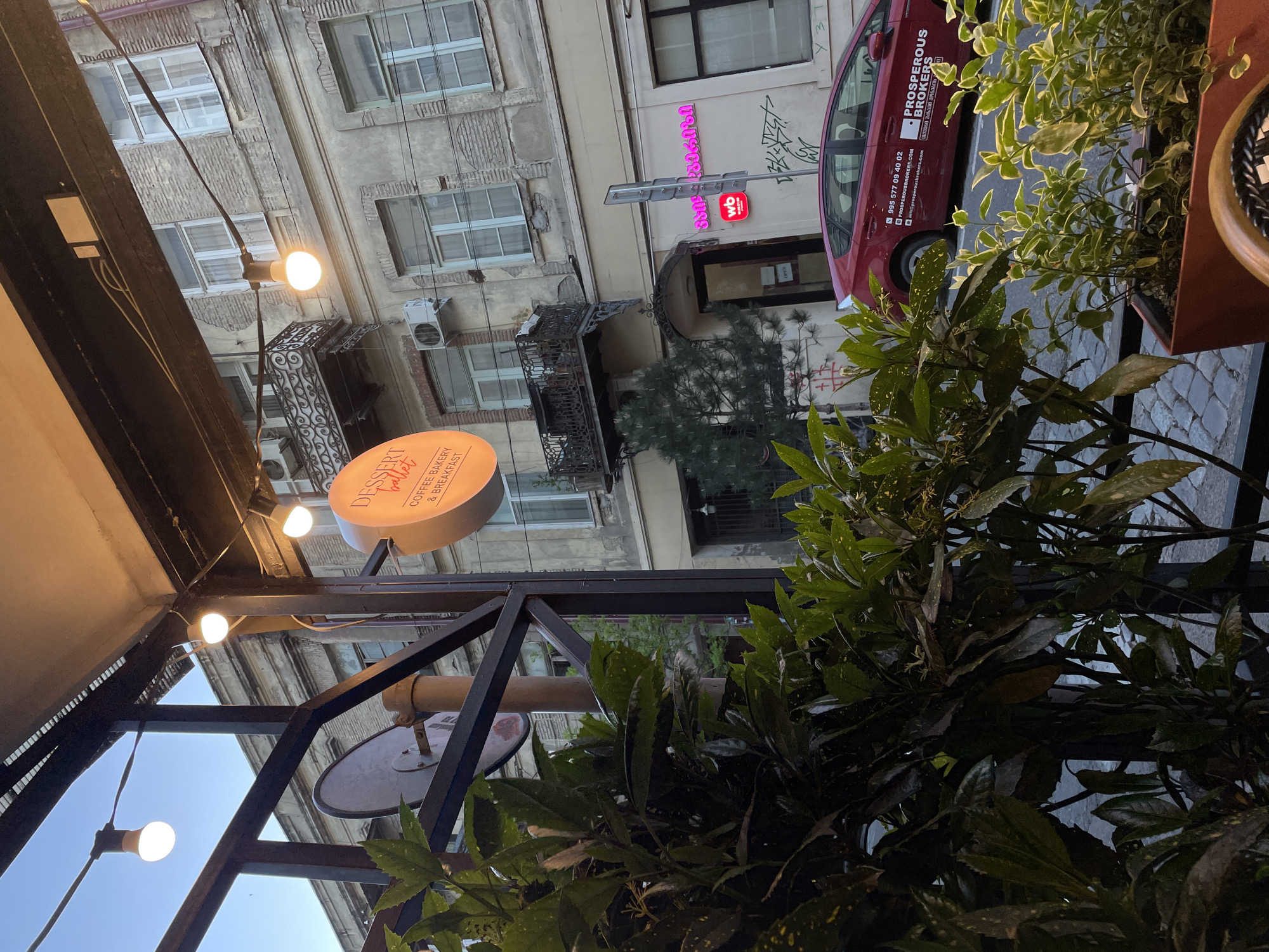 This image presents an inviting urban evening scene, likely from a cafe patio in Tbilisi, Georgia. The perspective looks across a street towards old, distinctive buildings, bathed in the soft light of early evening or twilight.
In the foreground, a dark metal structure supports several warm, glowing string lights, creating an intimate atmosphere. Lush green potted plants, possibly providing privacy or decoration, occupy the right side of the frame. A prominent round sign, with an orange hue, is visible on the left, displaying the text "DESSERT ballet COFFEE BAKERY & BREAKFAST," identifying the establishment.
Across a narrow, presumably cobbled street, the facades of aged multi-story buildings stand out. These structures feature numerous windows and some ornate iron balconies, with air conditioning units affixed to their exteriors. A red car is parked along the street, bearing the branding "PROSPEROUS BROKERS," a phone number "995 577 09 40 02," and the website "PROSPEROUSBROKERS.COM," along with corresponding Georgian text. Further down the street, a building displays a bright pink neon sign with Georgian characters and a "wb" logo.
The overall setting suggests a vibrant yet relaxed urban environment, characterized by traditional architecture and modern street elements. The glowing lights and plants create a cozy ambiance against the backdrop of the city.