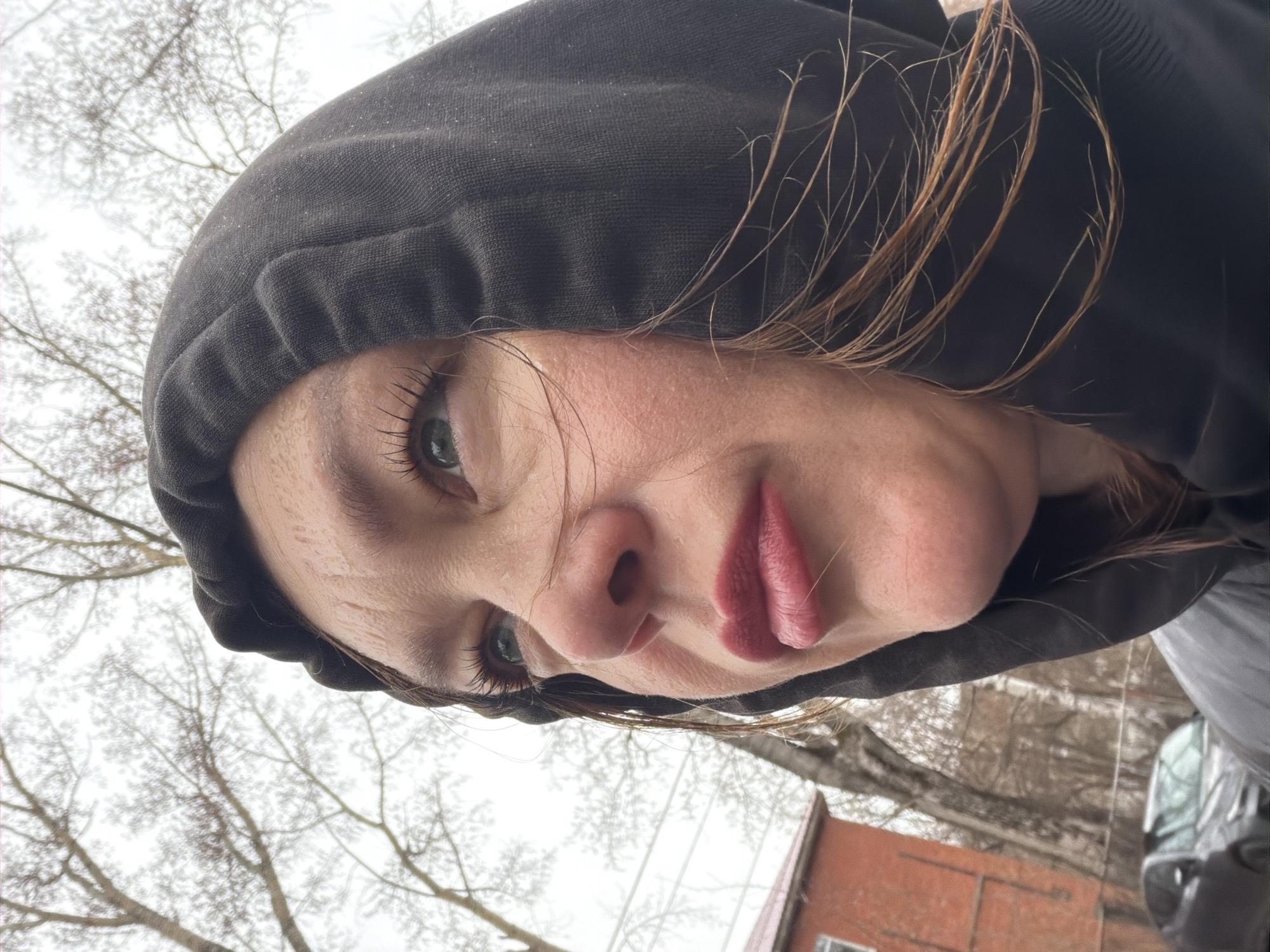 A selfie-style image shows a woman wearing a black hooded sweatshirt. The woman is fair-skinned with light-colored eyes and full lips. Stray strands of light brown hair are visible around her face. The photo is taken at an angle, with the camera looking slightly up at the woman's face.
In the background, bare trees are visible against a gray sky, suggesting a cold or winter season. In the lower portion of the background, a red brick building is partially visible, and to the right, a dark-colored car is also partially in view.
The location is identified as Chaikovsky, Russia.