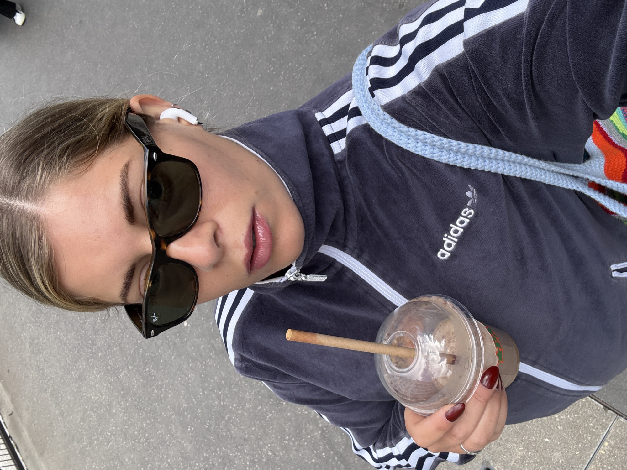 The image is a selfie taken from a high angle, showing a young woman wearing sunglasses, a dark-colored Adidas tracksuit jacket with white stripes, and an earpiece in her left ear. She holds a clear plastic cup containing a drink with a straw in her right hand. The Adidas logo is visible on the jacket. A light blue knitted bag strap is on her shoulder. The background appears to be a paved area, possibly a sidewalk. Given the style of the clothing and the drink, it is likely a candid snapshot taken in an urban environment. There is no information to confirm the location as Paris, France.