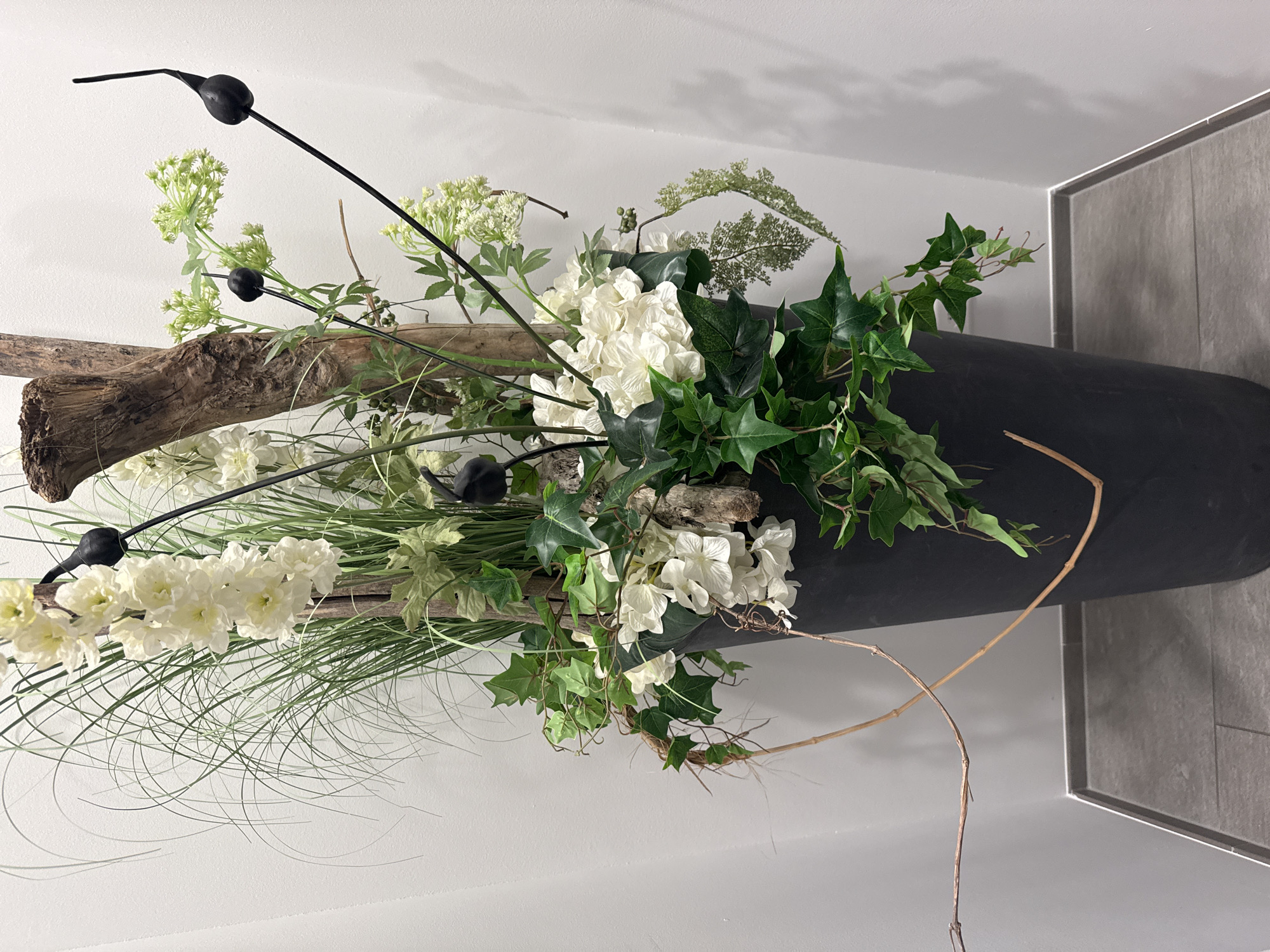 In Nendeln, Liechtenstein, a tall, dark gray planter holds an arrangement of artificial greenery and white flowers. The planter, which tapers downwards, is positioned against a plain white wall, with a gray tiled floor visible at its base. The floral arrangement includes long, wispy green grass-like elements, white clustered flowers resembling hydrangeas or delphiniums, and ivy with dark green leaves. A piece of rough, weathered wood serves as a central structural element. Black decorative spikes, each topped with a bulbous form, protrude from the arrangement. Thin, dry branches extend outwards from the planter and across the wall. The lighting is even, casting soft shadows on the wall.