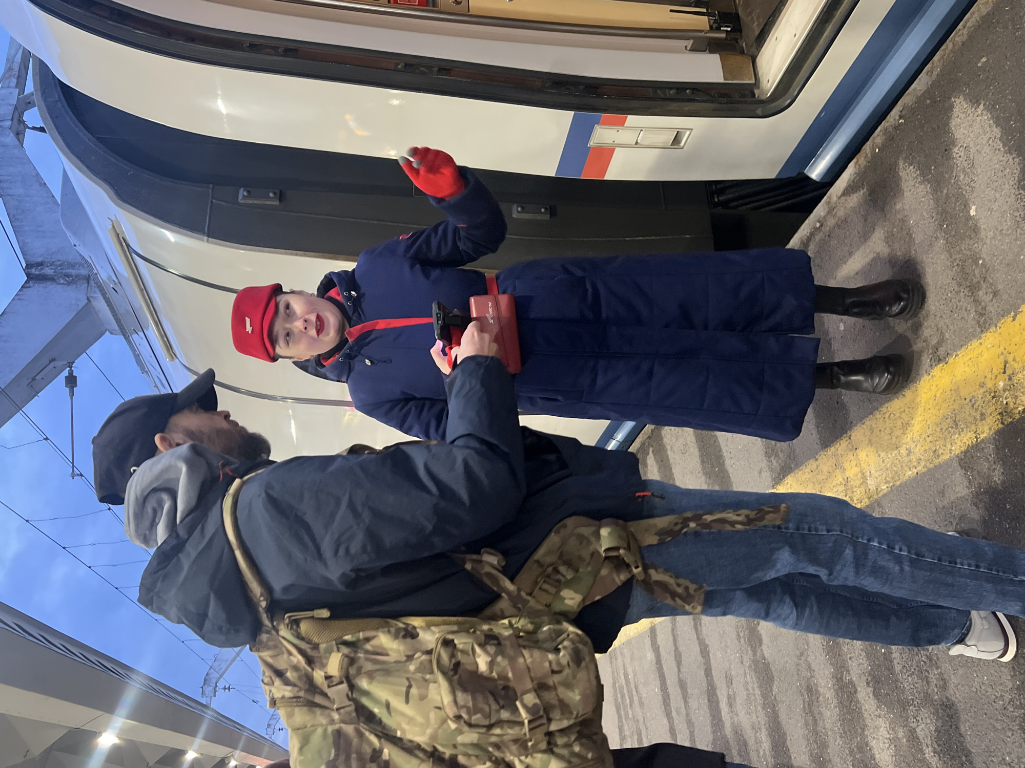 A train conductor in a blue uniform and red hat and gloves stands on a train platform next to a train. She is interacting with a man in jeans, a navy jacket, and a camouflage backpack. The platform is marked with a yellow line. The location is Saint Petersburg, Russia.