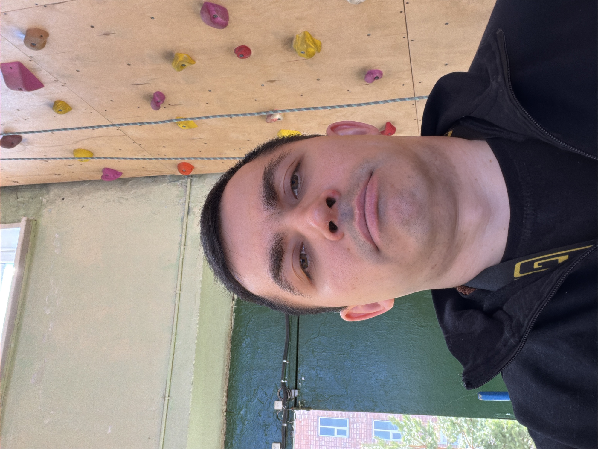 A selfie of a man in front of an indoor climbing wall is shown. The climbing wall has various colored holds. The man is wearing a black jacket. Outside the building is a glimpse of another building with light blue windows and green trees. Although the city of Izhevsk, Russia, is provided as context, there are no identifiable visual elements in the image that definitively confirm its location in that specific city.