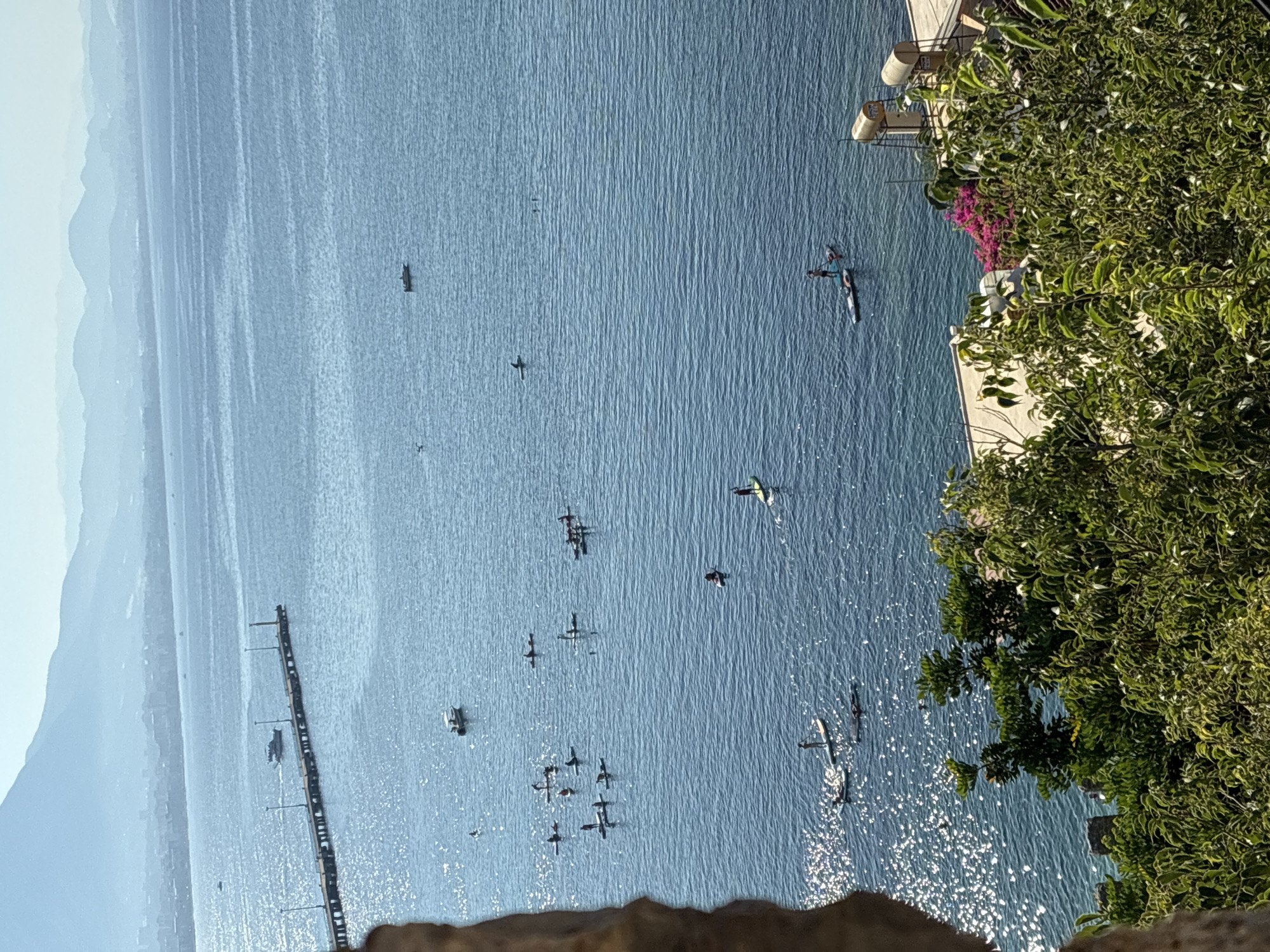 The image shows a scenic view of the Mediterranean Sea in Alanya, Turkey, as seen from a high vantage point. In the foreground, lush green foliage partially obscures the view, with a glimpse of a stone wall on the left. The middle ground is dominated by the shimmering blue sea, dotted with numerous people paddleboarding and a few small boats. A long pier extends into the sea from the left, with a boat docked at its end and several lamp posts lining its length. In the distance, the coastline of Alanya is visible, with buildings stretching along the shore, and rolling hills and mountains forming a hazy backdrop under a clear blue sky. Two solar water heaters are visible on a building on the right side of the image, near the water's edge. The overall scene depicts a serene day at the sea, with recreational activities taking place.