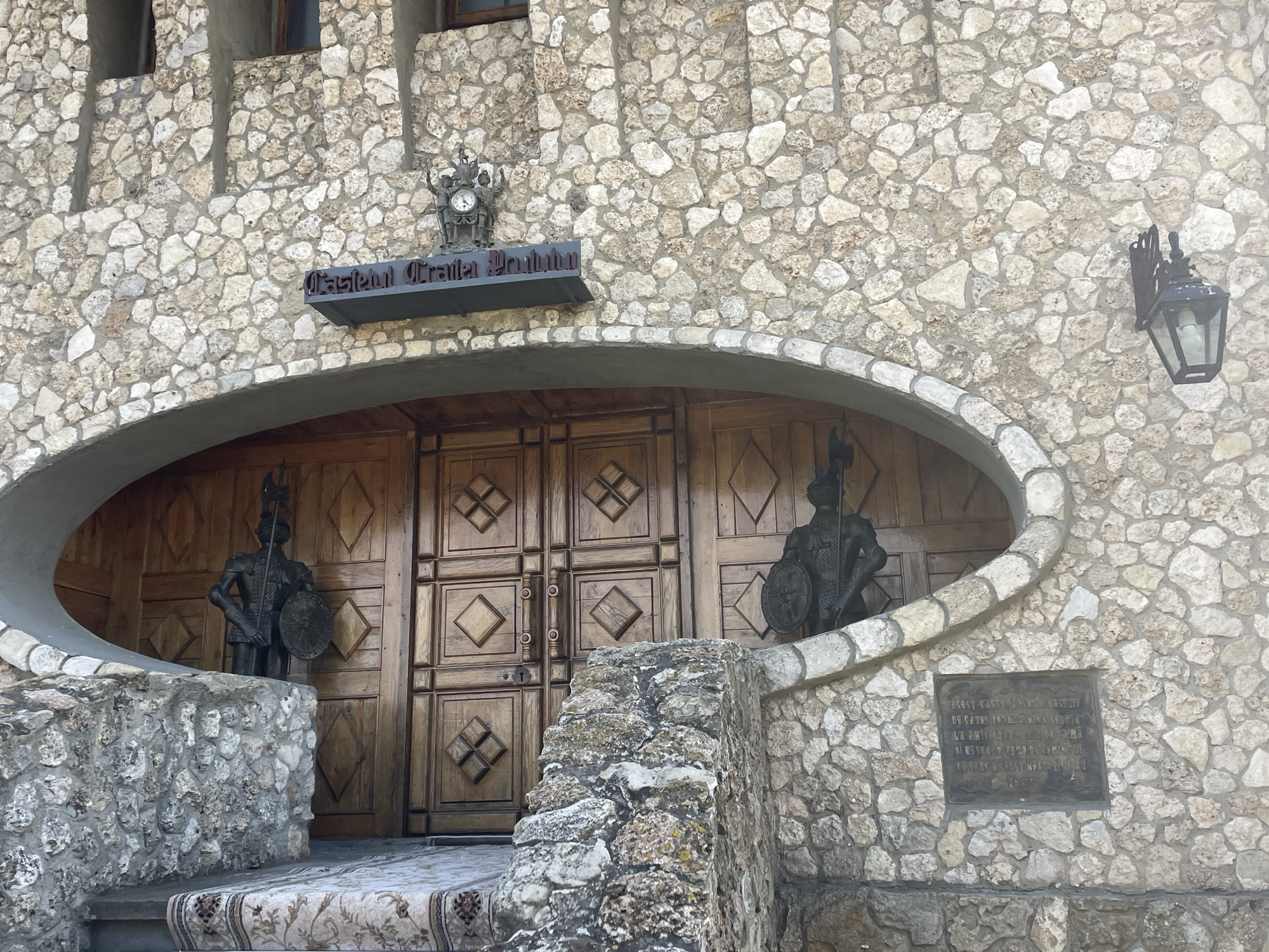 The image shows the entrance to the "Castelul Traniță" in Colibași, Moldova. The facade is constructed of rough-hewn stones, with a large, arched entryway that frames a set of imposing wooden double doors. Flanking the doors, within the archway, are two life-sized bronze statues of armored warriors holding shields and spears. Above the entrance, a sign with dark, ornate lettering reads "Castelul Traniță". To the left of the sign, a decorative clock face with figures adorns the stone wall. A black, wrought-iron lantern is mounted on the right side of the facade. A stone staircase leads up to the entrance, partially covered by a patterned carpet. A plaque with inscribed text is affixed to the stone wall to the right of the entrance. The overall impression is of a historical or themed building, with a rustic and somewhat imposing architectural style.