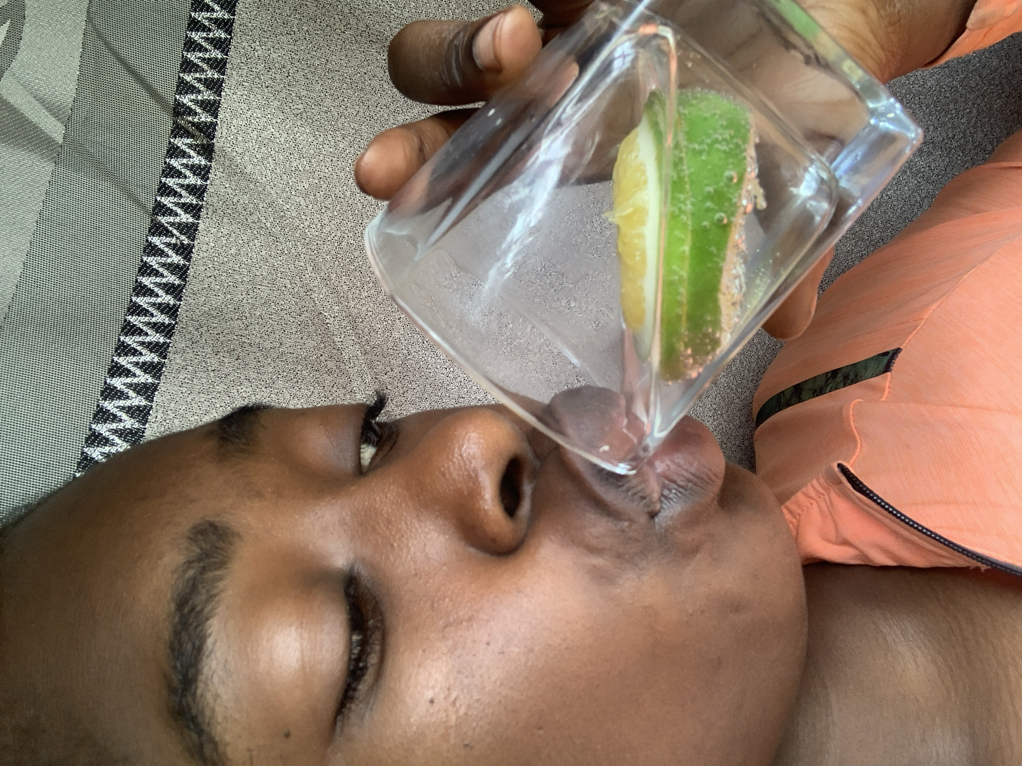 A close-up, overhead shot depicts a dark-skinned person drinking from a square glass containing a slice of lime and lemon. The person's eyes are closed, and the glass is tilted so the liquid is touching their lips. A hand is holding the glass. The person is lying on a grey and white patterned surface next to a peach-colored garment with black trim. The environment appears indoors. There is no indication of this photo being taken in Rafin Par, Nigeria.