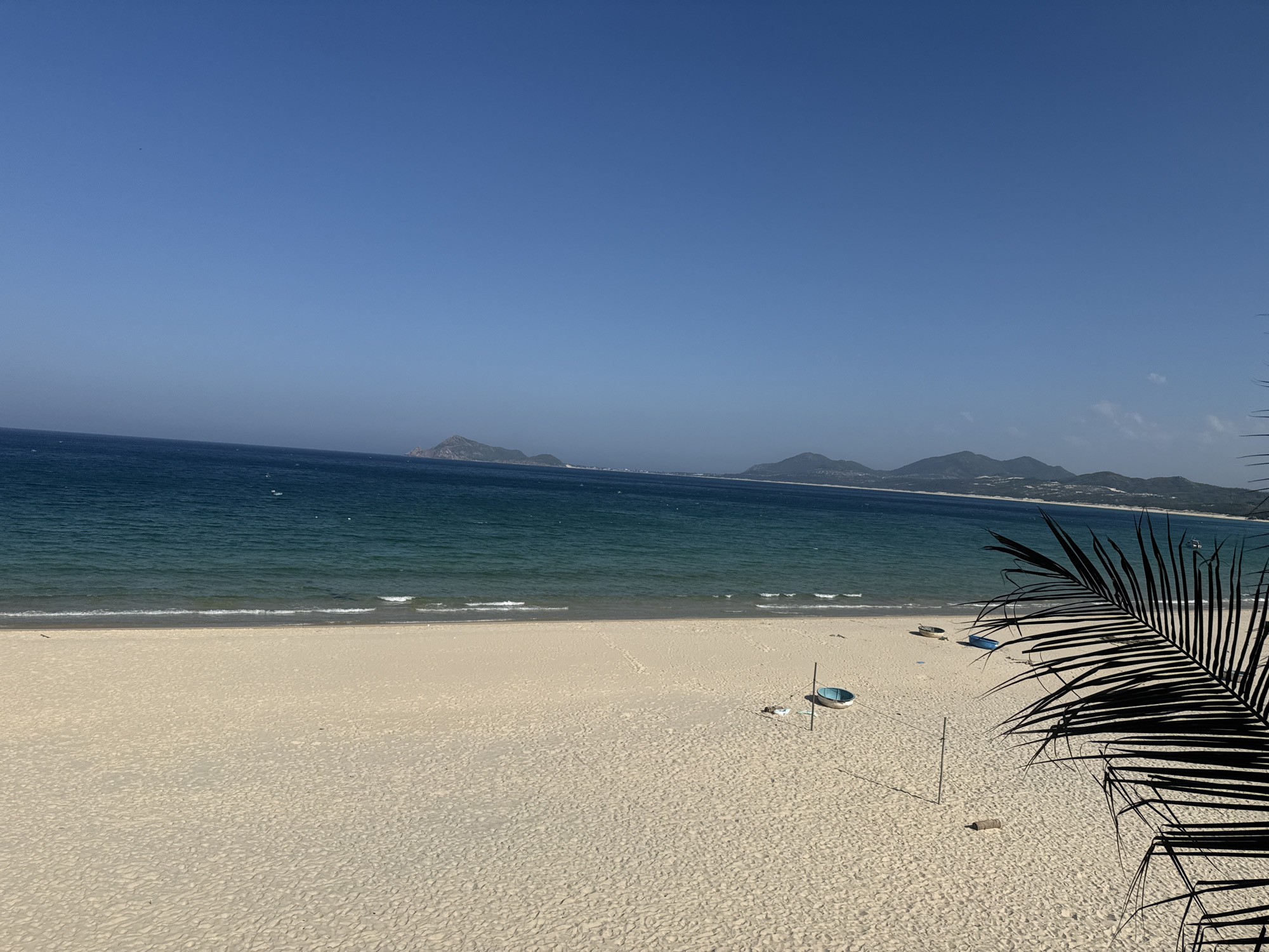 An aerial wide shot captures a tranquil beach scene under a clear blue sky. The vast expanse of the ocean, a deep teal, meets the light tan sand with gentle waves. Distant, hazy mountains form a rugged coastline on the horizon. On the sandy beach, a few small, colorful boats are visible, along with a single palm frond that partially frames the right side of the image, casting a shadow. The sand itself shows subtle textures and footprints, suggesting recent activity. The overall impression is one of a serene, natural coastal landscape.