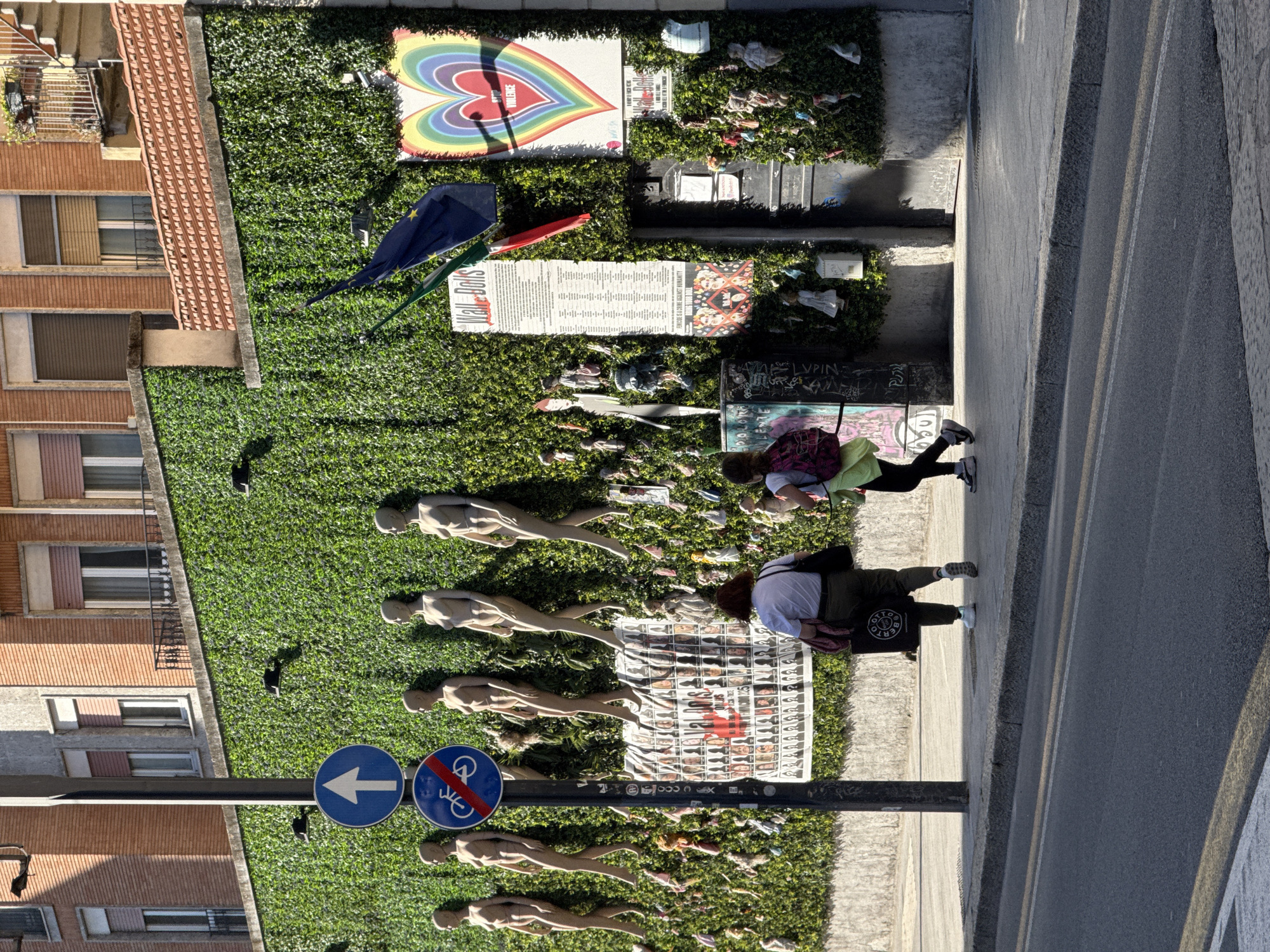 A street art installation, referred to as a 'Wall of Dolls', is displayed in Milan, Italy. The installation consists of several mannequin figures arranged on a faux grass backdrop. The figures are positioned horizontally along the street, some showing signs of wear. Beside them, an exhibition of various posters, signage, and artwork is present. The wall shows anti-violence images, including an image of a rainbow heart. There is also an image with multiple photos of violence victims.
Two women are observed examining the installation. One is wearing a black outfit and the other has on a green shirt and pink pants.
The location is urban with buildings adjacent to the sidewalk, and the street art is positioned along the edge of the road. The weather appears to be sunny, indicated by the strong shadows.
Also, traffic signals are set up, including a blue sign of an arrow pointing left and one showing a pedestrian with a bicycle.