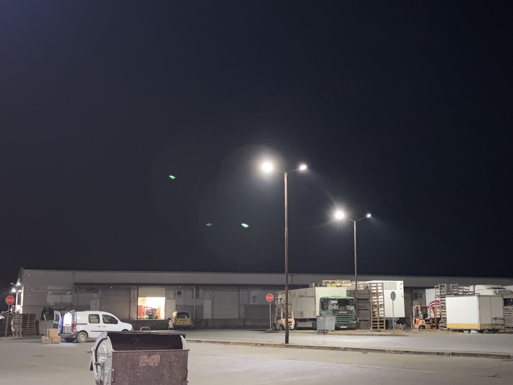 The image depicts an industrial area, likely a warehouse or distribution center, located in Plodovitovo, Bulgaria at night. The scene is illuminated by streetlights, with a long, low building forming the backdrop. Several delivery trucks and forklifts are parked near loading docks. A white van is parked in front of one of the open loading bays, with boxes and a person nearby. A large dumpster sits in the foreground. Stop signs are visible near the building. The sky is dark, with faint green artifacts possibly from camera.