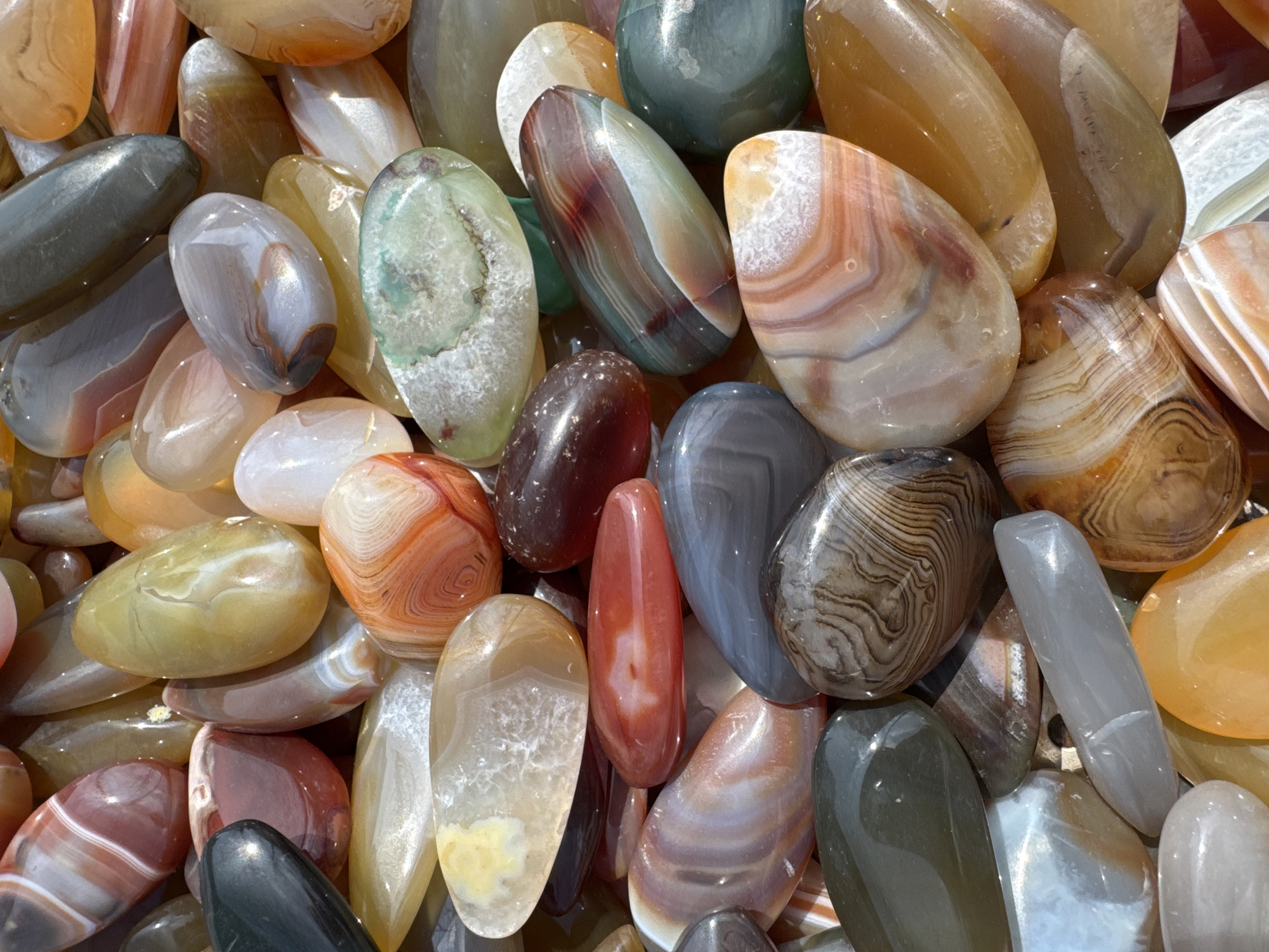 The image displays a dense aggregation of numerous tumbled and polished agate stones, filling the entire frame from a top-down perspective. These stones exhibit a wide range of natural colors, including vibrant reds, oranges, yellows, browns, greens, grays, whites, and dark, almost black tones. Many of the agates feature distinct banding patterns, swirling formations, and areas of translucency, with some stones appearing opaque or showcasing internal crystalline structures. All stones possess smooth, glossy surfaces that reflect ambient light. The scene is located in Wulingyuan, China. No individuals, explicit actions, or other environmental elements are visible within the depicted area.