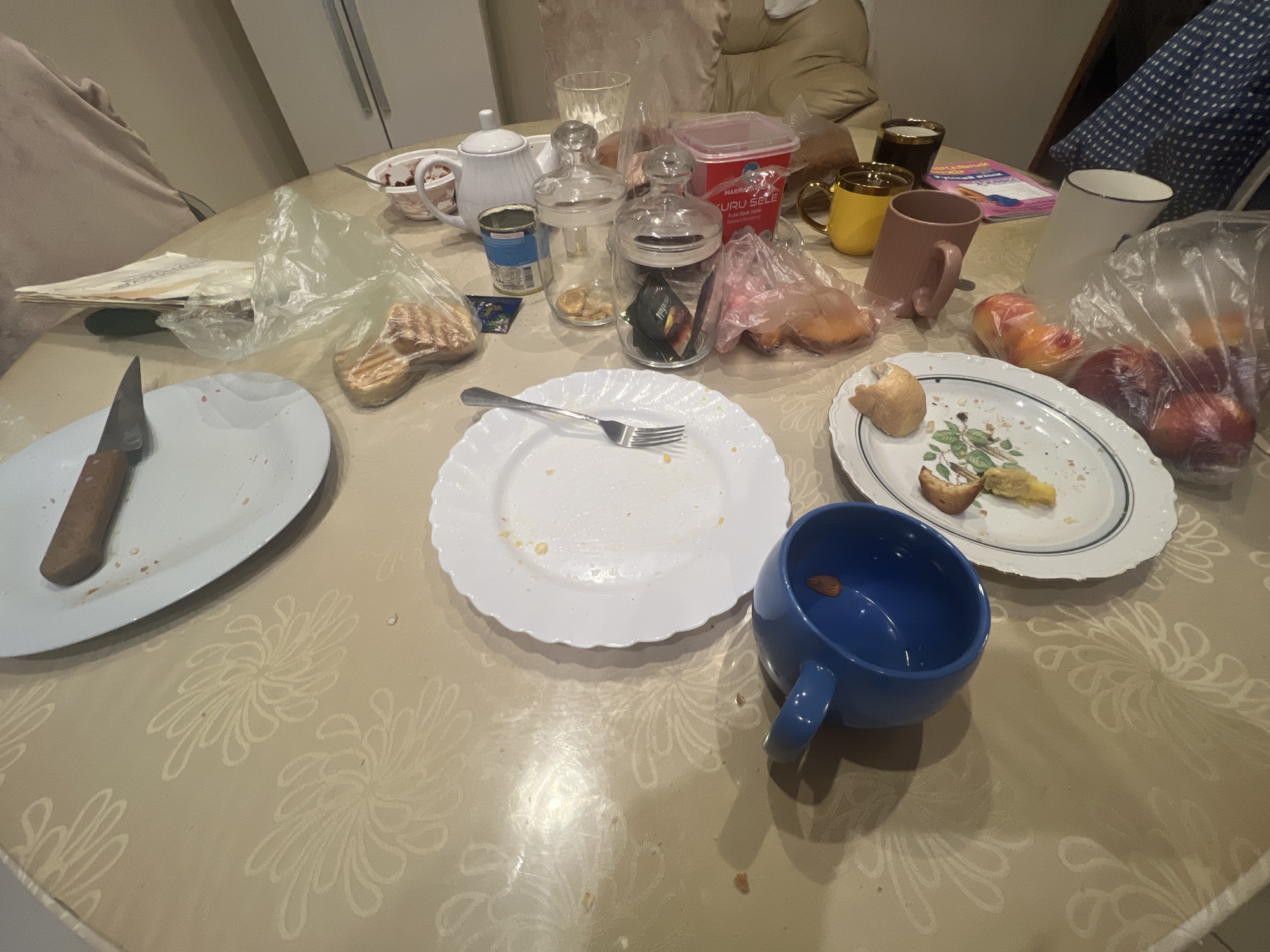 A round dining table, covered with a patterned tablecloth, is laden with remnants of a meal. The scene appears to be indoors, possibly in a home setting. A white teapot sits on the left, next to a small bowl containing a pinkish substance, possibly jam or compote. Several glass jars, some with lids, are scattered across the table, along with a red plastic container labeled "MARM " and "KURU SELE" in what appears to be Turkish script, suggesting the presence of some food product.
Several plates are visible, some clean and others with food scraps. A white oval plate on the left holds a knife with a wooden handle. A white round plate in the center has a fork resting on it and shows signs of being used. Another oval plate to the right features a few pieces of bread and some colorful food, possibly fruit. Several pieces of bread, wrapped in plastic, are also present.
Various mugs and cups are on the table: a yellow mug, a pink mug, a dark mug, and a tall white mug. A blue mug, positioned in the lower right of the frame, contains a single almond. In the background, a sofa or armchair is partially visible, suggesting a living or dining area. The lighting is relatively dim, hinting at an indoor setting without direct sunlight, possibly evening or a dimly lit room. The overall impression is that of a recently finished or ongoing meal, with items left on the table as if people are still present or have just departed. No specific time of day or weather conditions can be determined from the image, and no emotional cues are strongly evident beyond the casual nature of a meal.