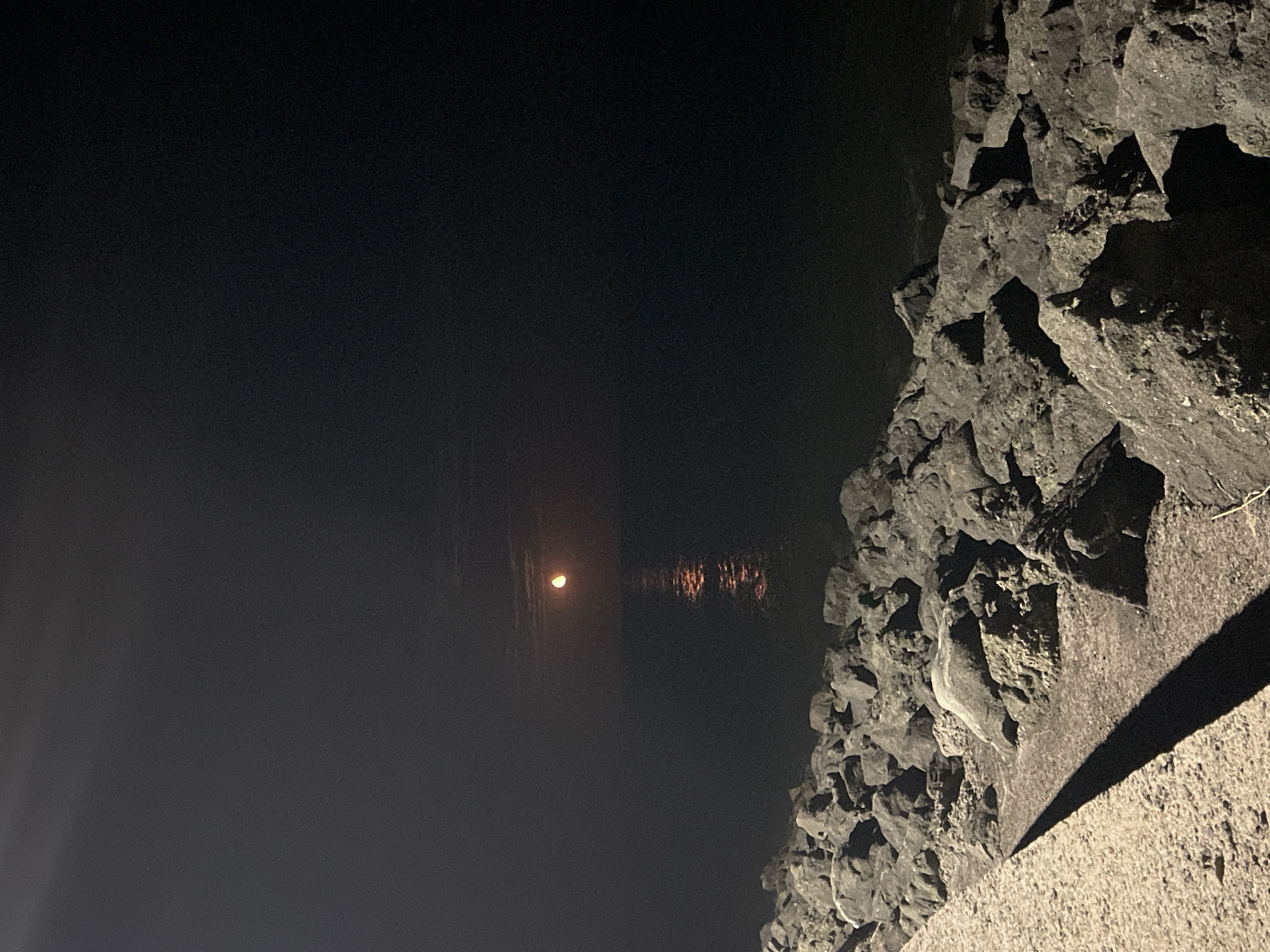 The image depicts a nighttime scene at a rocky shoreline, likely in Primorsko, Bulgaria. A full moon is visible in the dark sky, casting a shimmering reflection on the calm water. In the foreground, a concrete path leads to a dense arrangement of large, dark rocks that form a breakwater or natural barrier along the coast. The path is illuminated by an unseen light source, creating a long shadow cast by the rocks. The overall atmosphere is serene and tranquil, with the natural elements of moon, water, and stone dominating the composition. There are no people or animals visible in the image.