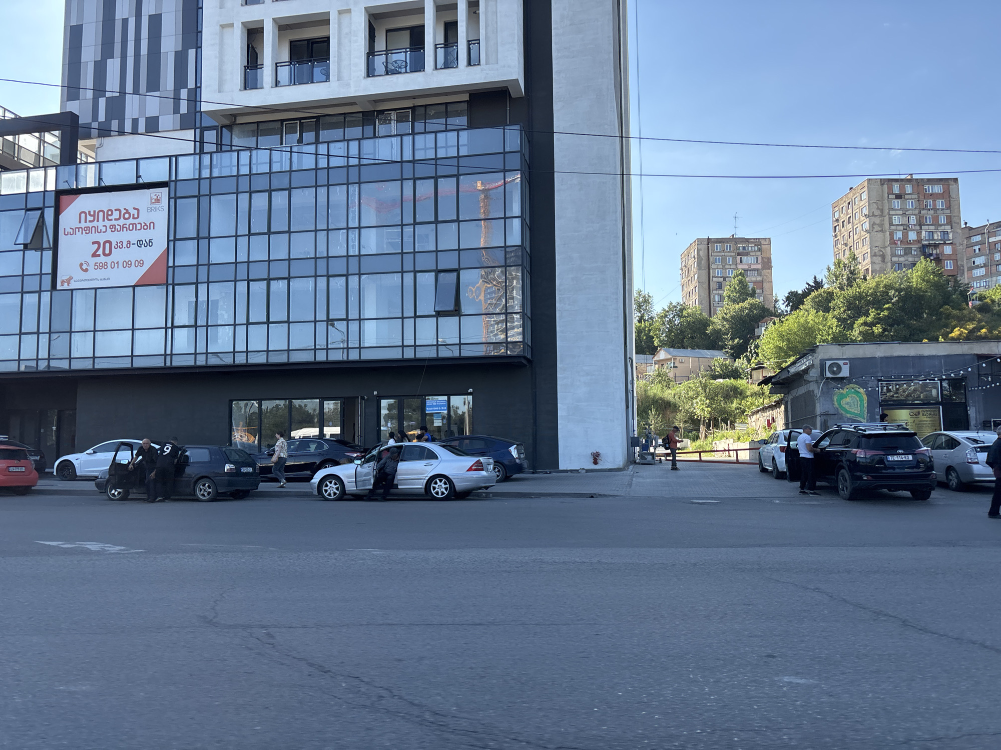 The image depicts a street scene in Tbilisi, Georgia. A multi-story modern building with glass facade dominates the left side of the frame. A red and white sign with Georgian text is displayed on the building's facade. Several cars are parked on the street in front of the building, and people are seen interacting near some of them. In the background, older residential buildings are visible on a hillside, along with trees and a clear blue sky. The foreground shows a paved street.