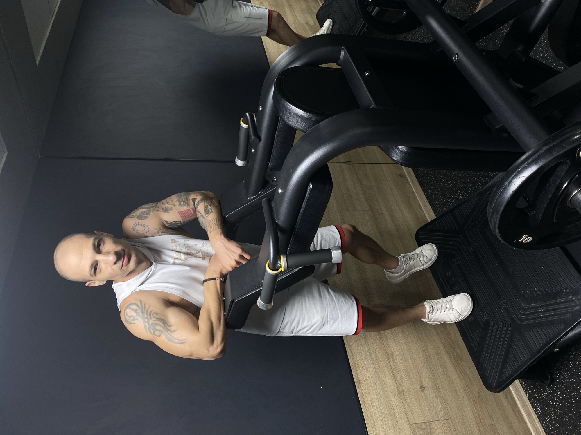 A muscular, bald, tattooed man wearing a white tank top and white shorts with a red trim is shown from an overhead perspective, operating a black leg press machine at a gym. The gym has a black accent wall and a light-colored wooden floor. The man is smiling and looking towards the camera. Plates are loaded on the machine. The location is likely in the city of Pleven, Bulgaria.