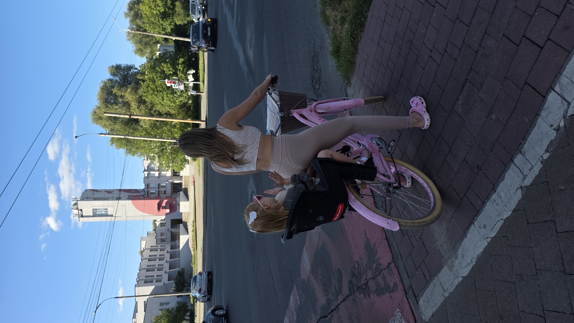 This image depicts a woman riding a pink bicycle on a brick-paved sidewalk. A young child is seated in a black child seat attached to the rear of the bicycle. The woman is wearing a white crop top and beige leggings, and the child is wearing a gray top and has pink sunglasses. They are on a street in Khmel'nyts'kyi, Ukraine, with a modern-looking building and a tall tower with a red design visible in the background. Cars are present on the road, and trees line the side of the street. The sky is blue with some clouds.
