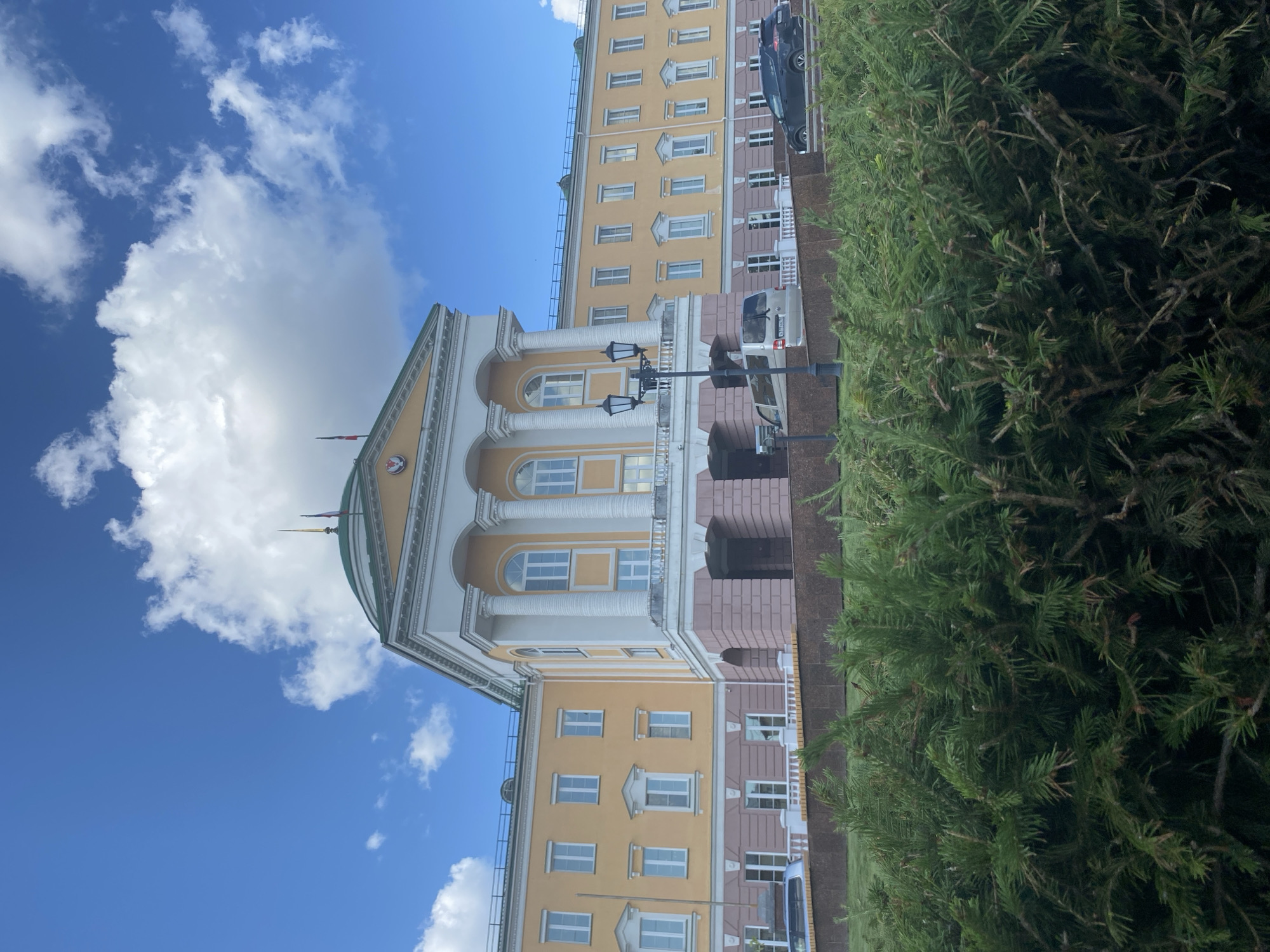 The image depicts the Udmurt State University, a prominent educational institution located in Izhevsk, Russia. The building is characterized by its yellow facade, classical columns, and a green dome. In the foreground, lush green bushes and trees partially obscure the lower portion of the university building and the surrounding grounds. Several vehicles, including a silver van and a black car, are visible parked on the street in front of the university. The sky above is a vibrant blue, dotted with fluffy white clouds, suggesting fair weather. The architectural style of the university suggests a formal and stately presence within the city.