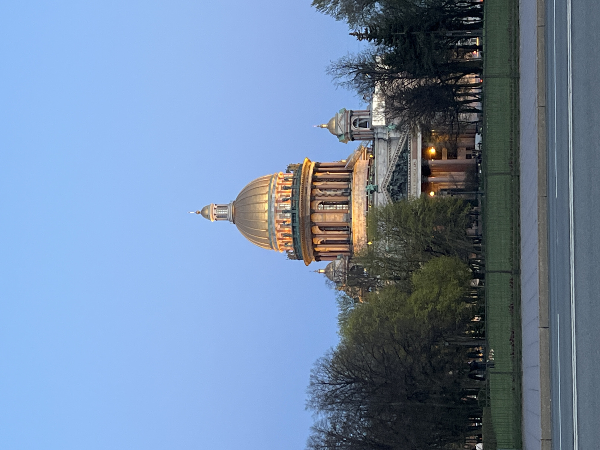 The image features Saint Isaac's Cathedral in Saint Petersburg, Russia. The cathedral, with its large golden dome, is centrally positioned, illuminated by lights, set against a twilight blue sky. Trees with varying degrees of foliage frame the cathedral. A green area, separated by a fence, sits next to an asphalt road with painted white lines. No people are visible in the scene.