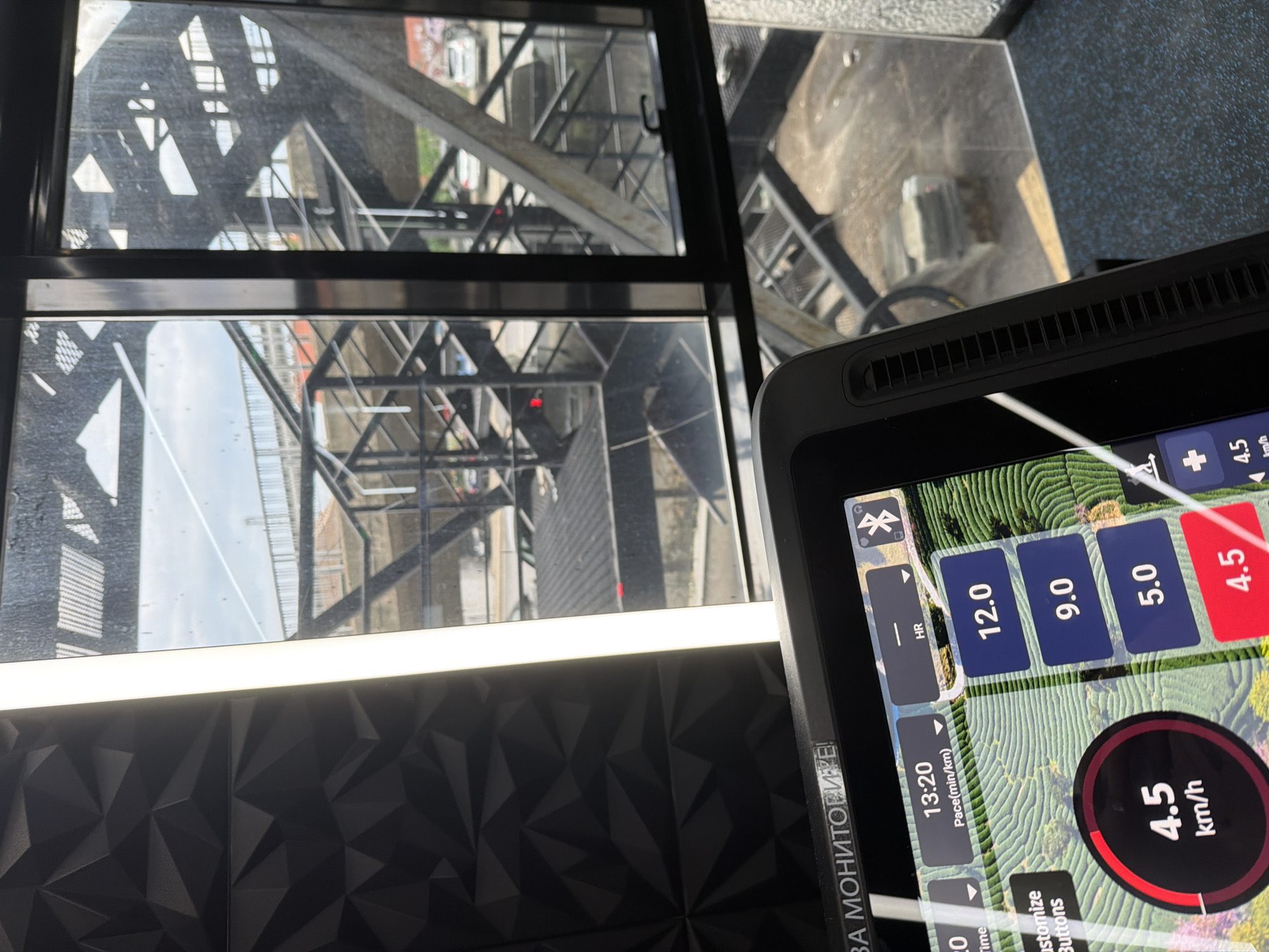 The image shows a close-up view of a treadmill's console with a reflection in the window behind it. The console displays various metrics such as "HR", "12.0", "9.0", "5.0", and "4.5 km/h". There is also a clock showing "13:20" and "Pace(min/km)". The background seen through the window appears to be an urban industrial area with metal structures and some buildings. The lighting suggests it is daytime, possibly afternoon, with a bright overcast sky. The treadmill is in focus, highlighting the digital display which suggests an exercise or workout is in progress. The reflection in the window shows a similar industrial landscape. The overall scene is functional and focuses on the technology of the exercise equipment. Visible text includes workout data and a Cyrillic inscription that appears to say "ЗА МОНИТОРИТЕ!" which translates to "FOR THE MONITORS!".