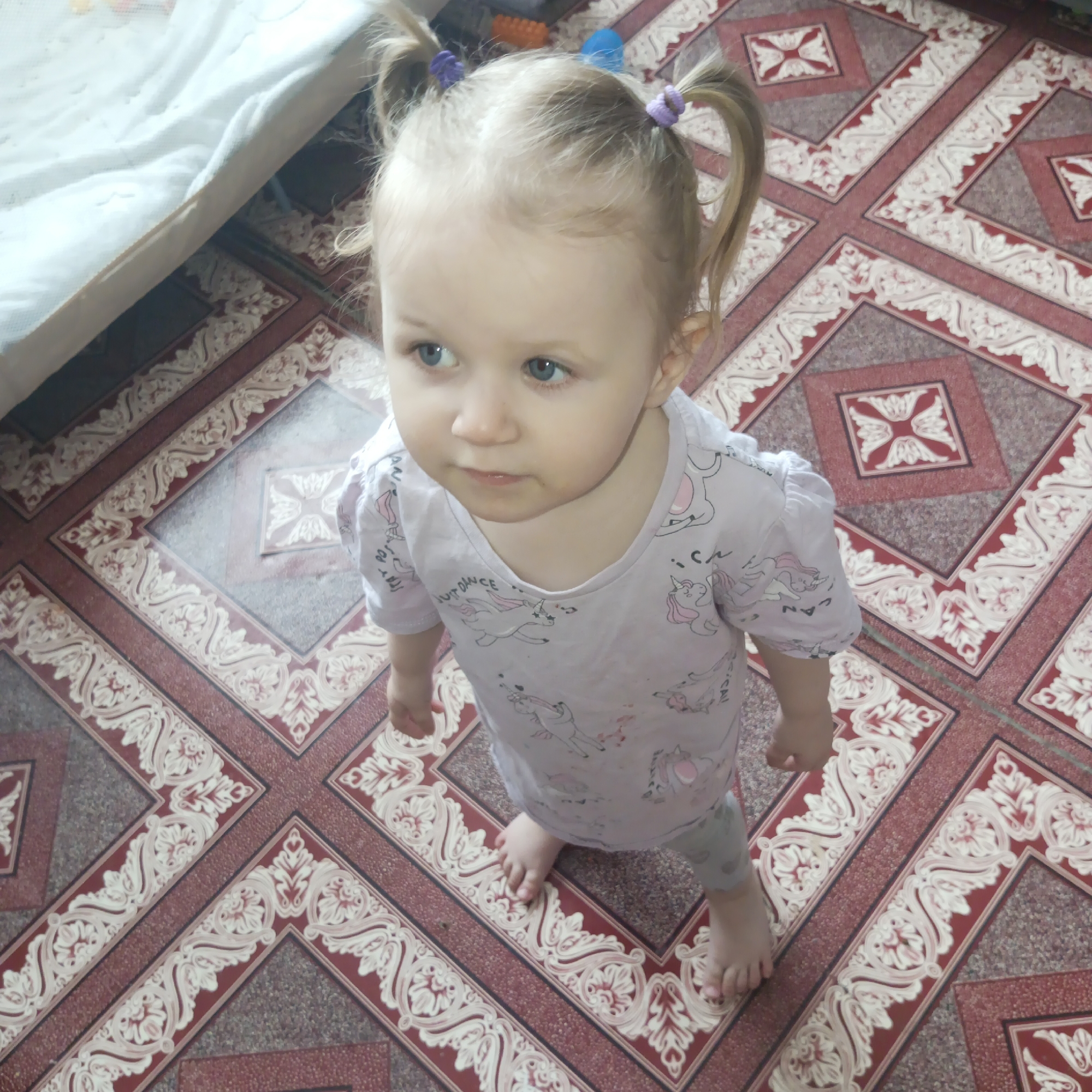 A fair-skinned, blonde-haired toddler with blue eyes stands on a patterned floor in what appears to be an indoor setting. The child is wearing a light purple T-shirt with unicorn prints and gray leggings. Her hair is styled into two pigtails, each secured with a purple and blue hair tie. The floor features a repeating pattern of squares with ornate designs in red, white, and gray. A portion of a bed or sofa covered with a white sheet is visible in the upper left corner of the image. Based on the available information, the city location of Chaikovsky, Russia cannot be determined.