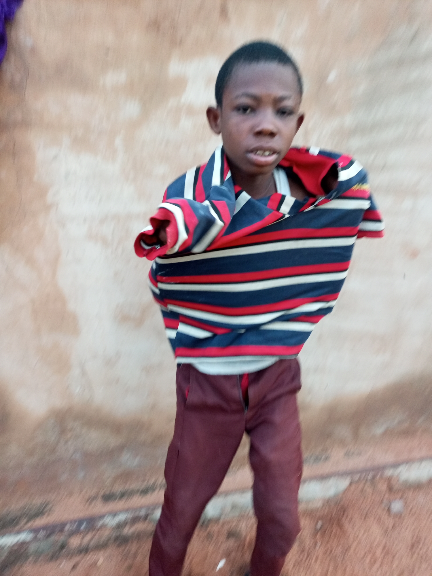 A young boy stands in front of a textured, beige wall. He is wearing a striped shirt with horizontal bands of navy blue, red, and white, which appears to be pulled down, revealing a white undershirt. He also wears dark maroon pants. The background wall has a rough, uneven surface with variations in color, suggesting it is made of stucco or plaster. The ground in the foreground is a mix of sand and dirt, with some debris visible. The lighting suggests it is daytime, possibly overcast, as there are no harsh shadows. The boy is looking towards the camera with a neutral expression. The image quality is moderate, with some motion blur, particularly in the lower half of the frame.