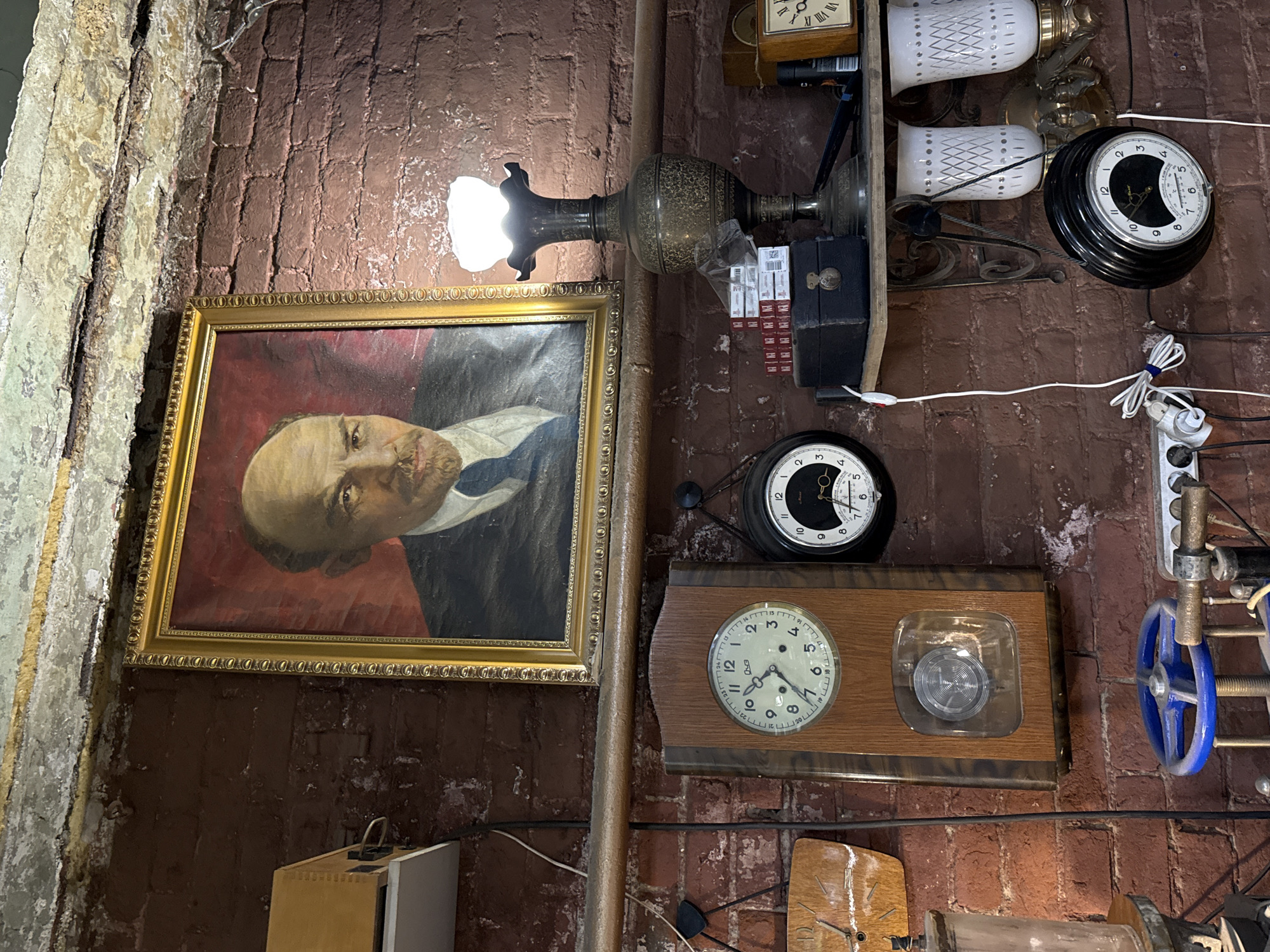 This is a medium shot, eye-level, indoor photograph of a brick wall adorned with various items, including a framed portrait of Vladimir Lenin, several clocks, and a lamp. The wall is constructed of dark red bricks with visible mortar. The ceiling above is cracked and aged, with peeling paint and exposed beams.
The most prominent item is a framed portrait of Lenin, positioned centrally on the wall. The portrait is a painting in a gold ornate frame. To the right of the portrait, a vintage lamp with an ornate metal base and a frosted glass shade is lit, casting a warm glow. Below the lamp and portrait, a wooden shelf holds several stacks of cigarette packs, a black box with a latch, and a vintage wooden box with a clock face.
To the left of the shelf, a large wooden-cased clock is mounted on the wall, featuring a round clock face with Roman numerals and a pendulum visible through a glass window at the bottom. To the right of this clock, a black circular clock is also mounted, with a white clock face and Roman numerals.
Further down the wall, a blue valve wheel is visible attached to some mechanical equipment. A power strip with several outlets and plugs is mounted low on the wall. Two white, decorative glass lampshades are mounted on the wall to the right of the shelf. The overall scene suggests an interior space with an eclectic collection of items, possibly a shop or a personal collection.
The photograph was taken in the city of Perm, Russia, specifically at the "Museum of Contemporary History of Perm Territory." The image captures an exhibit that displays historical artifacts and memorabilia.
