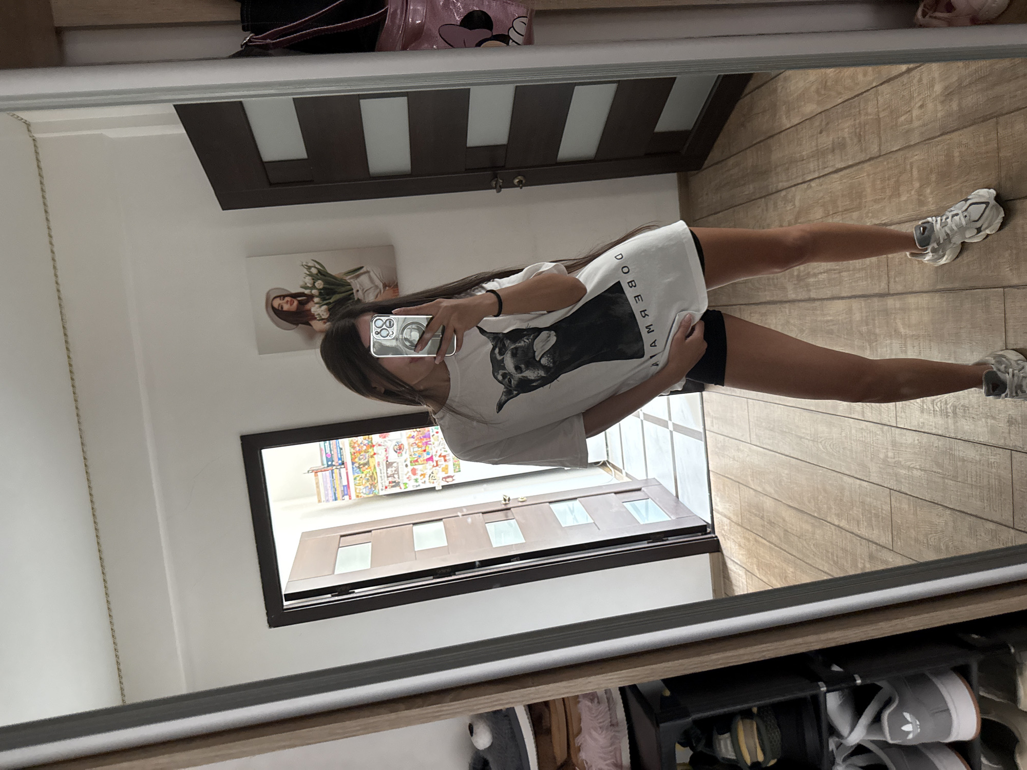 A young woman stands in a room, taking a mirror selfie. She is wearing a white t-shirt with a dog graphic and black shorts, and her hair is long and dark. She is holding a phone with a ring light in her right hand, and her left hand is tucked into the waistband of her shorts. The room has wooden floors and a tall mirror. In the background, there are two doors, one open and one closed, and a shelf unit with shoes. On the wall above the closed door, there is a framed picture of a woman with flowers. The scene appears to be a residential interior, likely an apartment or house.