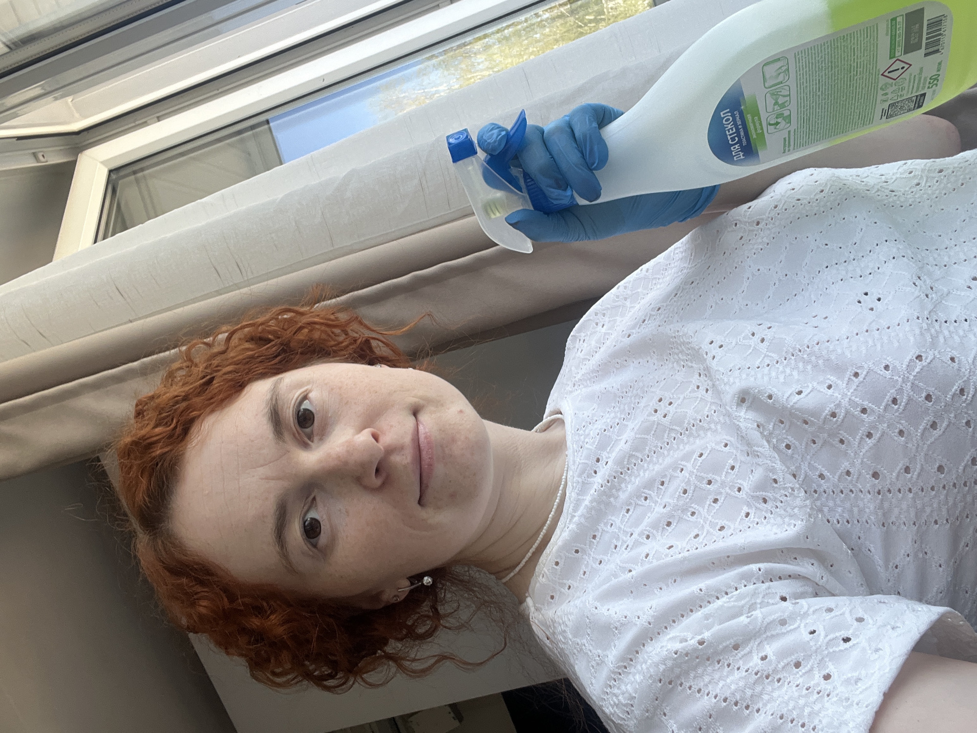 A woman with red curly hair and fair skin is holding a bottle of glass cleaner with blue gloves in front of a window. The bottle has Russian text on the label. The woman is wearing a white, lacy blouse and a pearl necklace. The window appears to be a skylight or a similar window design. The location is assumed to be in the city of Chaikovsky, Russia, based on the text on the cleaning product.
