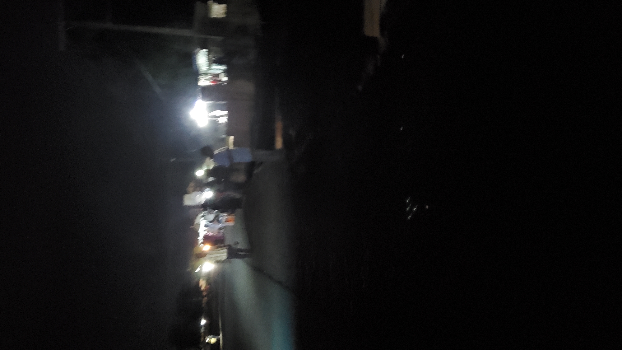 The image depicts a nighttime scene on a street in Jalingo, Nigeria. The lighting is dim and artificial, with several bright light sources illuminating parts of the scene. Several people are visible, appearing to be gathered or walking along the street. Some individuals are standing in groups, while others are in motion. There are structures visible in the background, possibly shops or market stalls, which are also illuminated by lights. The road surface is dark and appears to be paved. The overall impression is of a busy street scene at night, with people interacting and likely engaging in commerce or social activities.