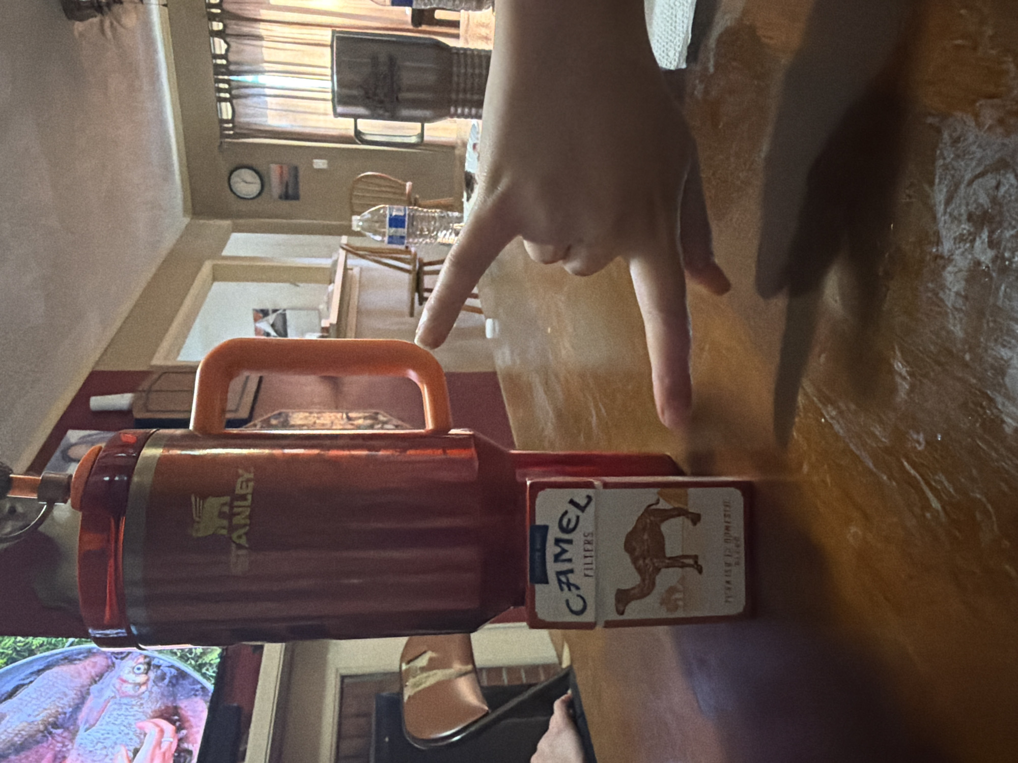 A close-up, eye-level shot shows a red Stanley brand thermal cup with an orange handle and a pack of Camel Filters cigarettes resting on a wooden table. A person's hands are positioned in the foreground, with one hand appearing to make a "shaka" sign. In the background, a television screen displays an image of fish, and further back, a clock, curtains, and a ceiling fan are visible, suggesting an indoor residential setting. The scene is lit by artificial and natural light, casting shadows on the table. The image is undated and lacks specific location details for Virginia Beach, USA.