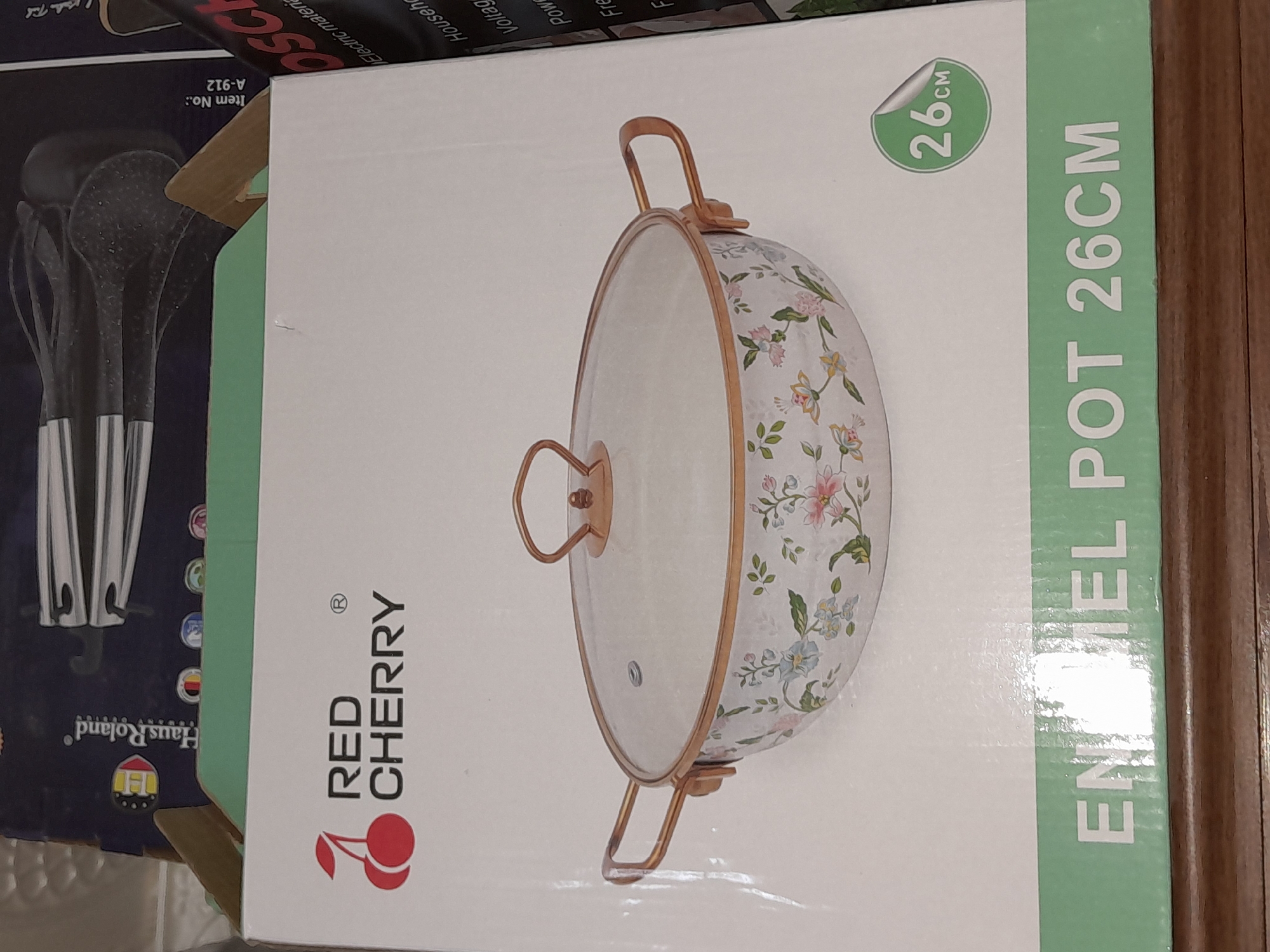 The image shows the packaging of a "RED CHERRY ENAMEL POT 26CM". The box is primarily white with a floral pattern on the pot depicted on the front. A green stripe at the bottom of the box displays the text "ENAMEL POT 26CM". A circular sticker indicating "26CM" is visible on the right side of the box. In the background, partially visible are other boxes, one with "Haus Roland" branding and another with "OSCH" branding. The scene appears to be an indoor setting, possibly a retail environment or storage area, with a wooden surface visible at the bottom of the image. There are no people or animals present in the image. The location of Uchquduq, Uzbekistan, is not discernible from the visual information provided in the image.