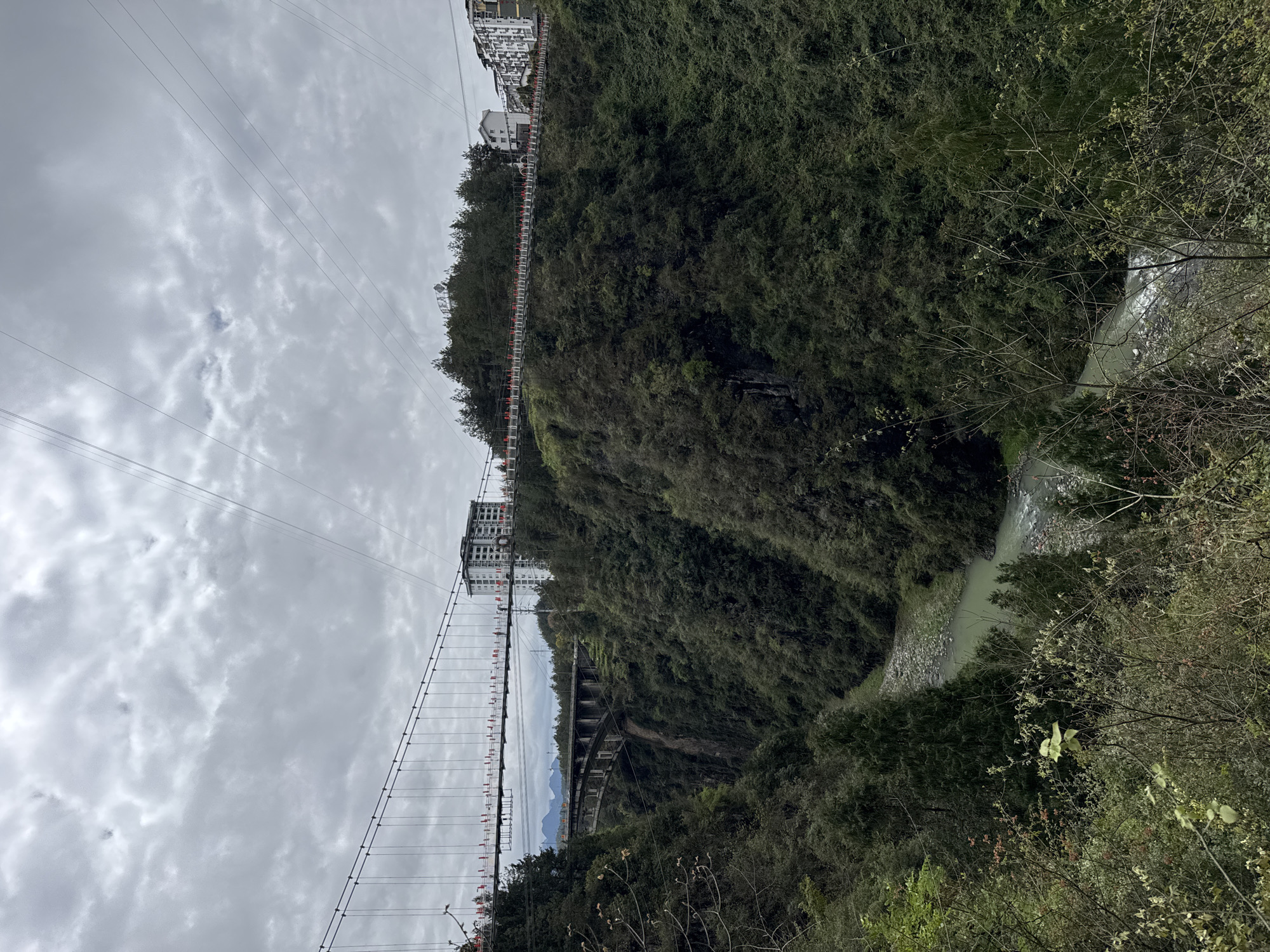 The image depicts a gorge with a river at the bottom, flanked by steep, green hills. Two bridges span the gorge, one a suspension bridge with red accents, and the other a lower, concrete arch bridge. Buildings are visible atop the hill on the left side of the image. Overhead power lines stretch across the cloudy sky. The river appears milky green in color. The scene is located in Qianjiang, China.