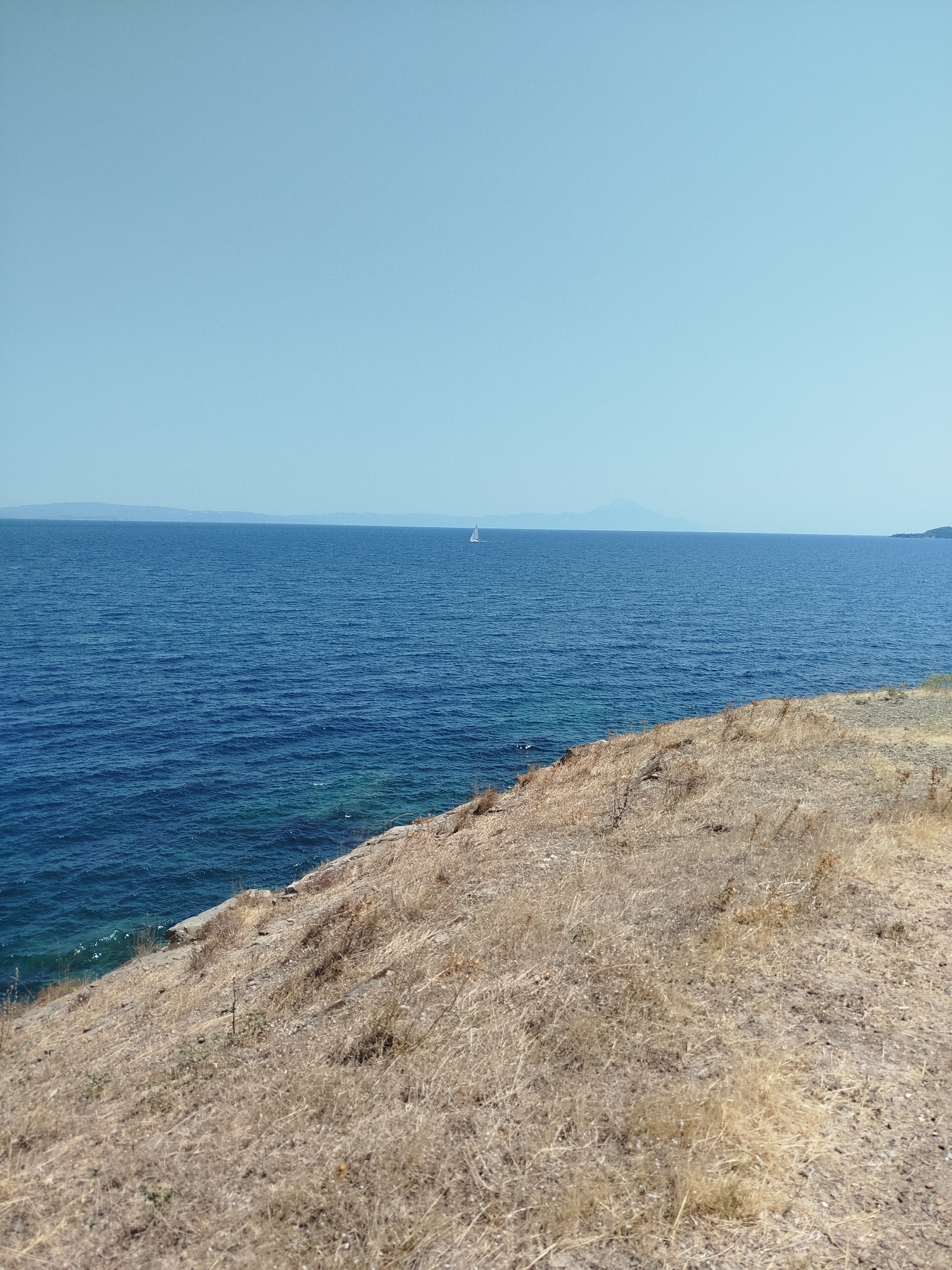 This is a wide-angle, eye-level shot of a coastal landscape in Sithonia, Greece. The image depicts a vast expanse of deep blue sea meeting a pale blue sky, with a thin line of distant mountains on the horizon. A small white sailboat is visible in the middle of the sea. In the foreground, a dry, grassy hillside slopes down towards the water. The vegetation is predominantly brown and yellow, suggesting a dry, sunny climate. The ground appears rocky and uneven. The overall impression is one of a serene, natural setting.