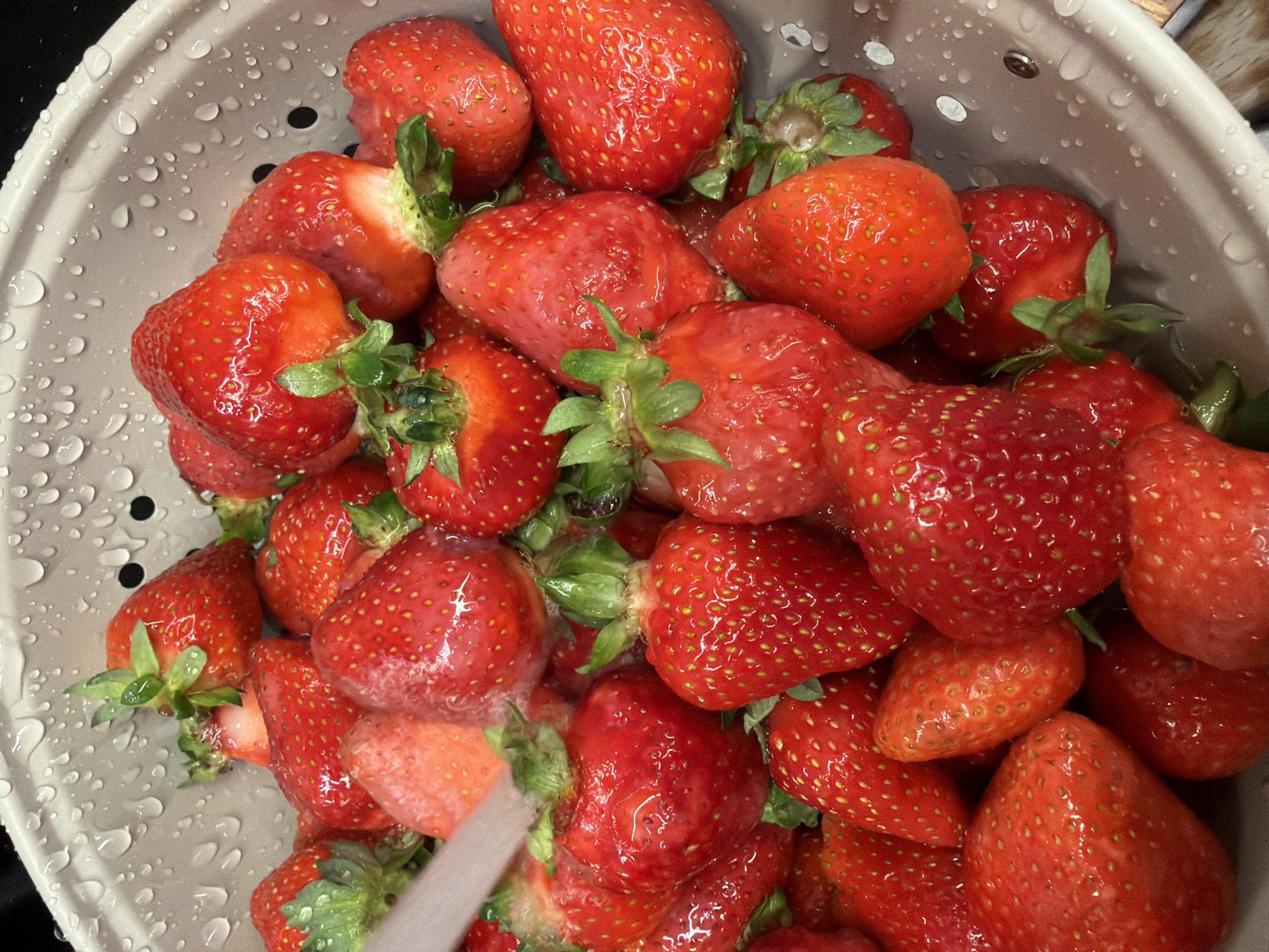 The image shows a colander filled with fresh, wet strawberries. The colander appears to be plastic or metal with drainage holes. The strawberries are red and ripe, with green stems. There are water droplets on the strawberries and the colander, indicating they have recently been washed. The image does not contain any information that would allow me to identify the location as Yerevan, Armenia.