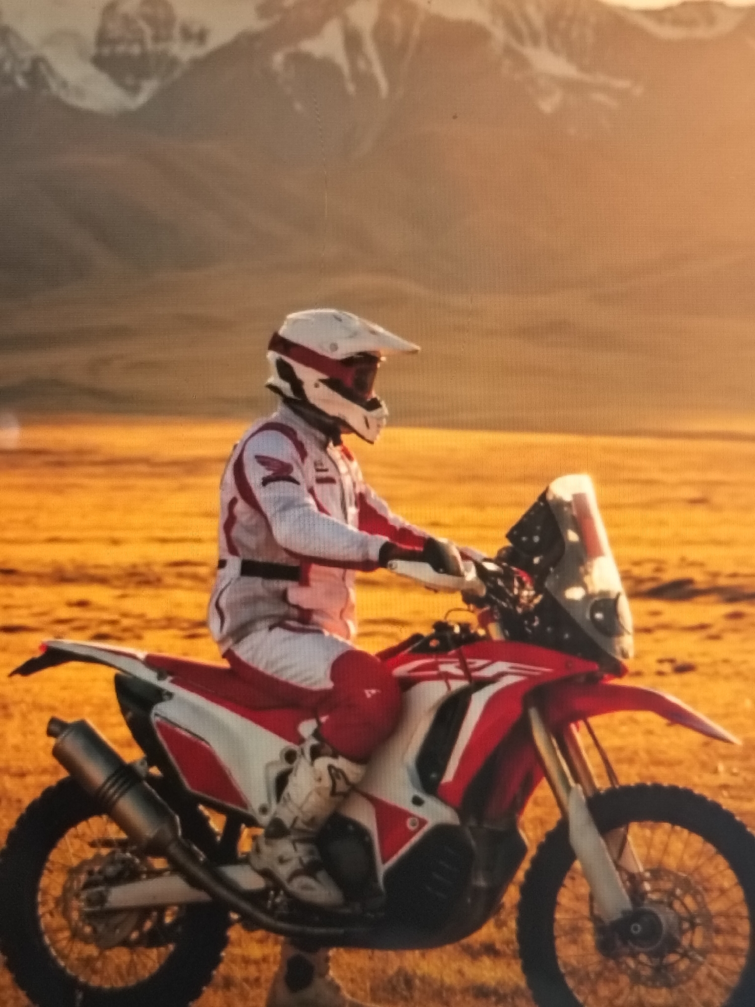 In Payagaboh, Indonesia, a person wearing a white and red racing suit and helmet is seated on a red and white dirt bike. The motorcycle has a prominent windshield and is parked on a dry, golden, and barren landscape. In the background, a range of imposing mountains with snow-capped peaks are visible against a hazy, warm-toned sky, suggesting either sunrise or sunset. The scene appears to be outdoors, in a remote, possibly desert-like terrain with sparse vegetation.