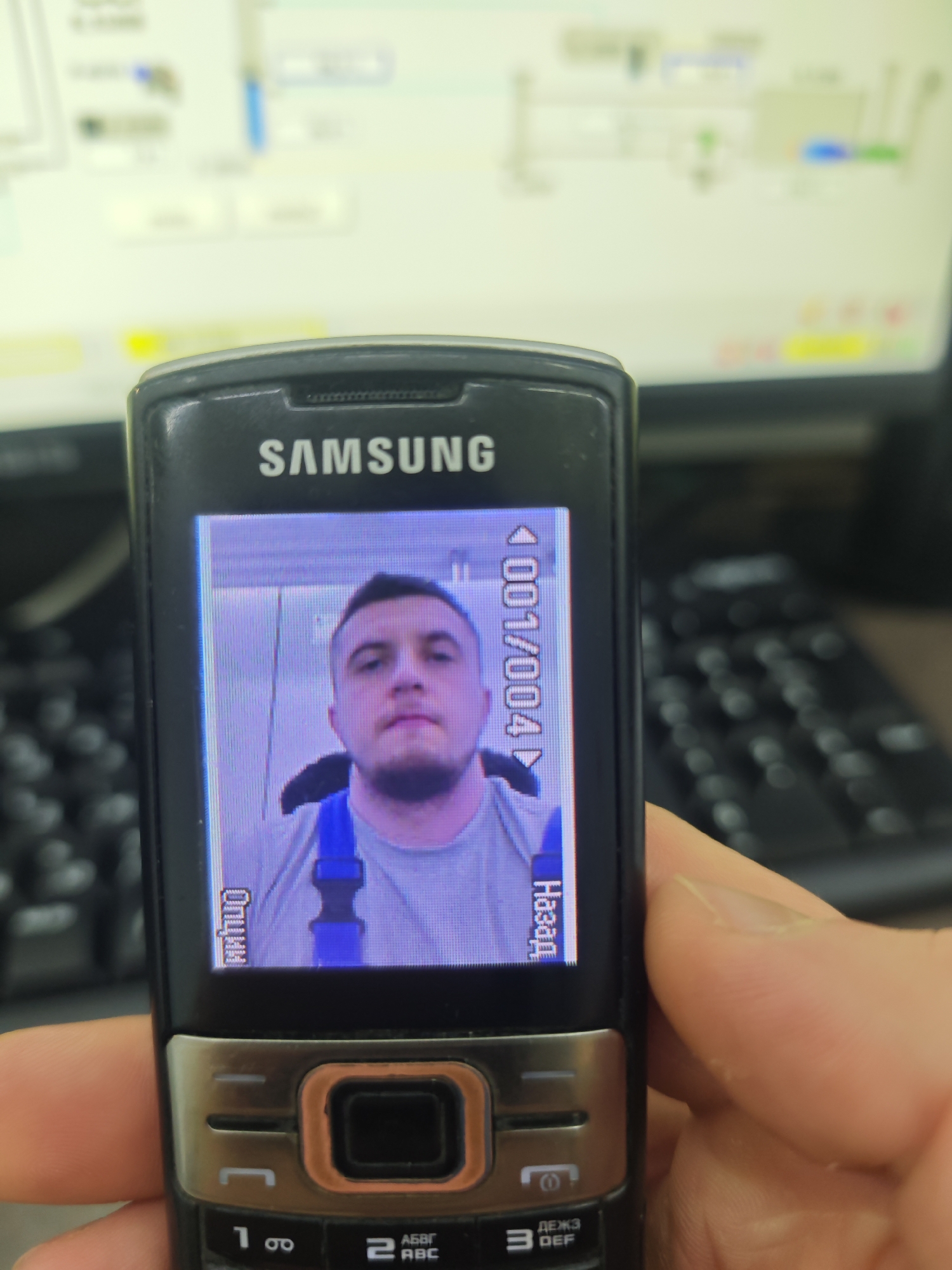 A Samsung feature phone is being held in a hand. The phone's screen displays an image of a man wearing a grey t-shirt and blue overalls. The phone's screen also displays "<001/004>" and the Russian words "Дальше" (Further) and "Назад" (Back). The phone has a silver and black finish and a numeric keypad. The background is blurred, but shows a computer monitor and keyboard. The location is unknown.