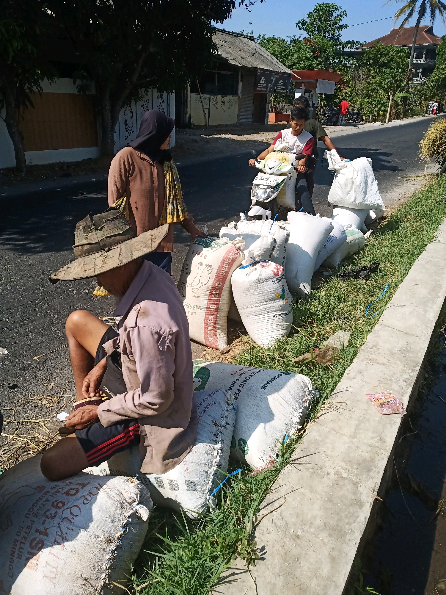 A street scene in Garut, Indonesia, shows an elderly man sitting on a large white sack, wearing a straw hat, a light purple shirt, and dark shorts with red stripes. He is holding something in his hands, possibly a small tool or object. Behind him, a woman wearing a brown hijab and a patterned scarf is standing. In the background, a young man on a scooter is being pushed by another person. Several large white sacks are lined up along the side of the road, some on the asphalt and others on the grassy embankment next to a concrete channel. In the further background, buildings and trees are visible under a clear blue sky.