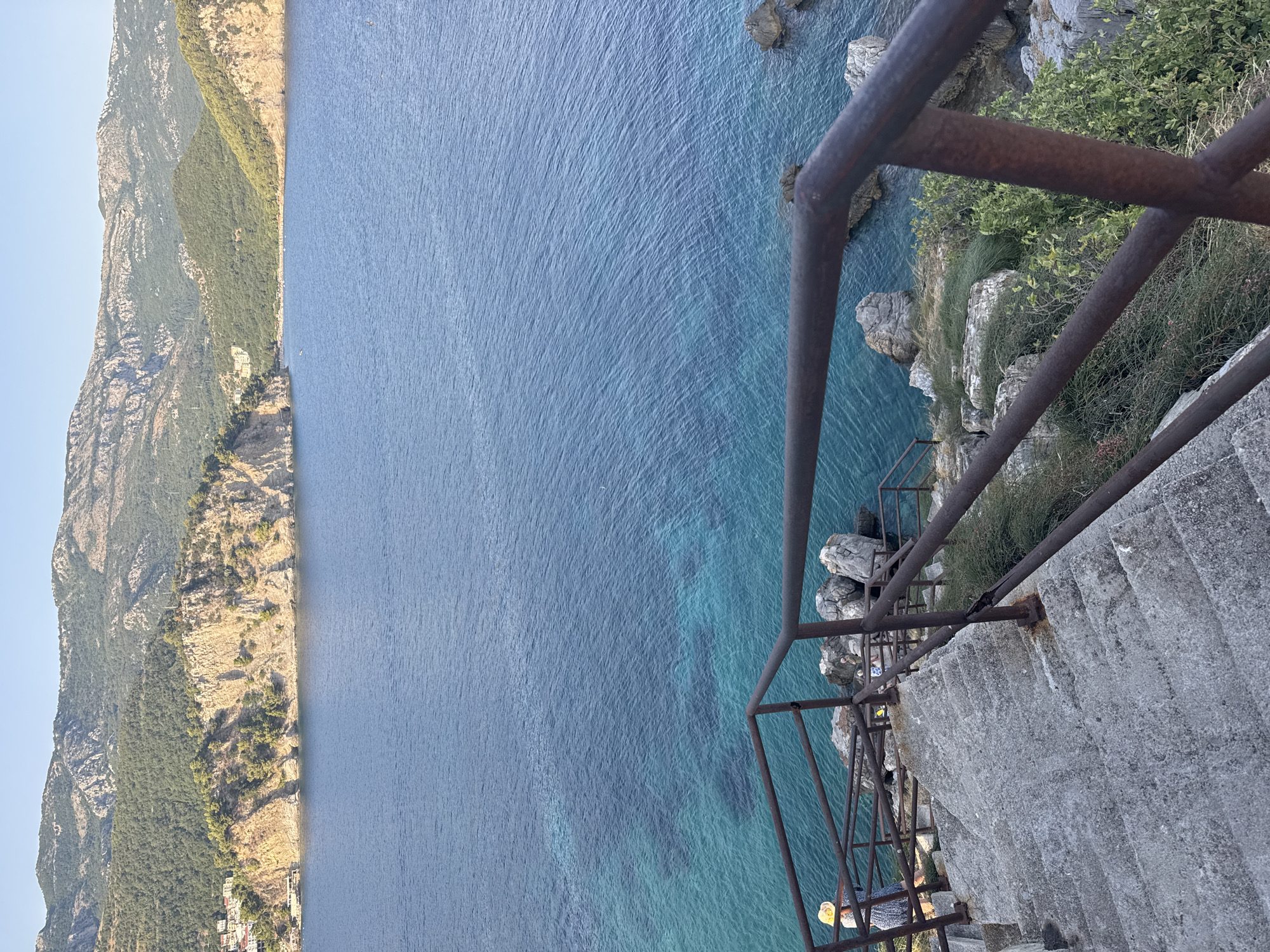 A high-angle, medium shot captures a scene of a stone staircase descending towards a beautiful blue body of water, likely in Petrovac centar, Montenegro. The staircase is worn and made of concrete, with a rusted metal railing running along its side. At the bottom of the visible stairs, a person wearing a yellow sun hat and a blue and white patterned dress is standing, facing away from the camera and towards the water. The water itself is a striking turquoise near the shore, gradually deepening to a rich blue further out. Rocky outcrops dot the coastline. In the background, dramatic, tree-covered mountains rise behind the water, with a scattering of white buildings visible on the slopes of the leftmost mountain. The sky above is a clear, pale blue. The overall impression is one of a serene, picturesque coastal landscape.