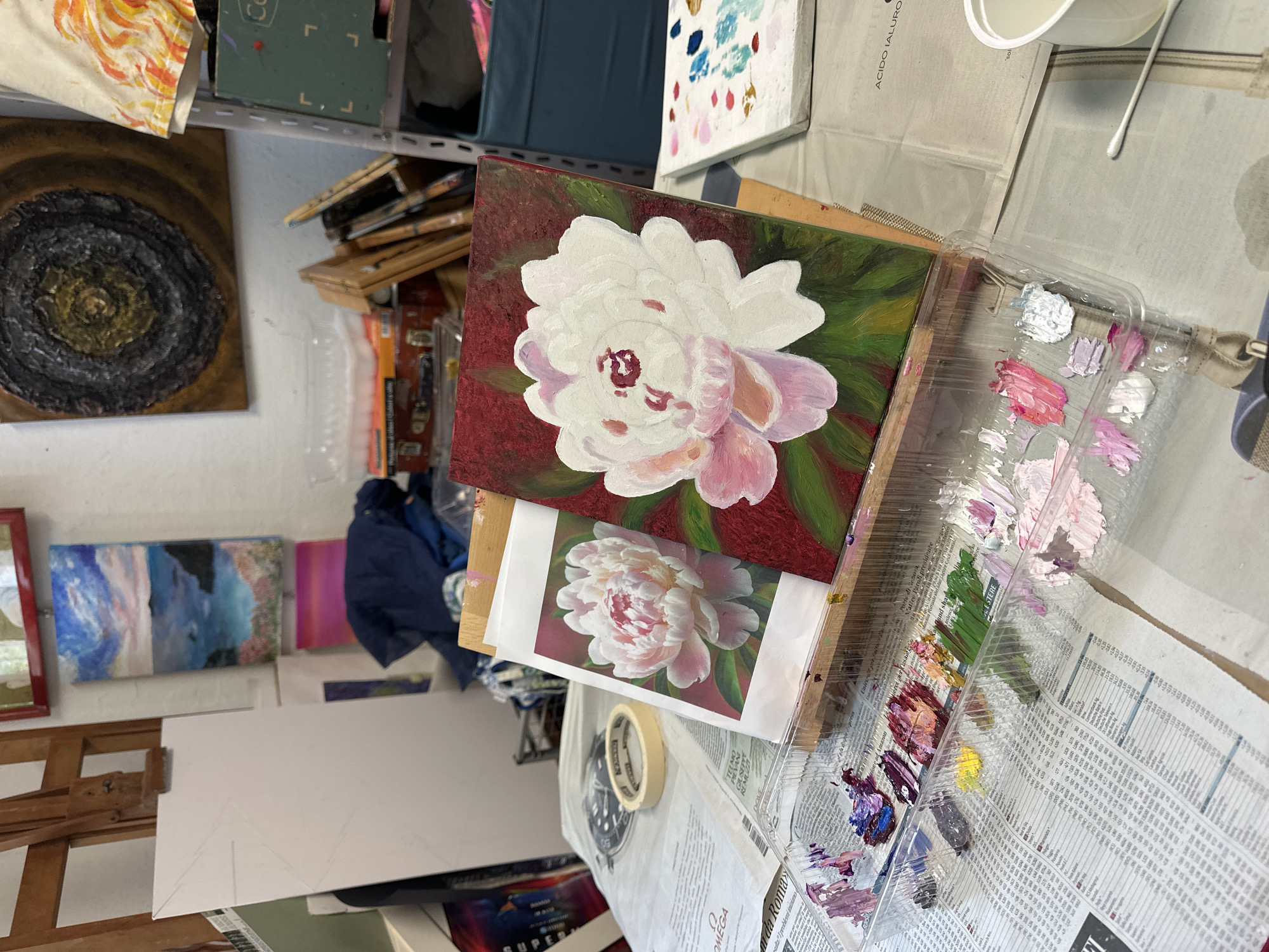 The image depicts an art studio setting with art supplies and ongoing projects. In the foreground, a clear plastic palette holds various dabs of paint in shades of pink, purple, white, yellow, and green, resting on a newspaper. Next to it, a painting on a canvas featuring a white and pink peony against a dark red background is propped on a wooden easel. Beside the painting is a printed reference image of a similar peony. In the background, other artworks are visible, including a landscape painting of a coastline and a textured circular abstract piece. Shelving units hold art supplies, including paint tubes and boxes. The overall environment suggests a creative workspace, likely in an artist's studio in Milan, Italy, given the presence of the newspaper with Italian text. There are no people visible in the image.